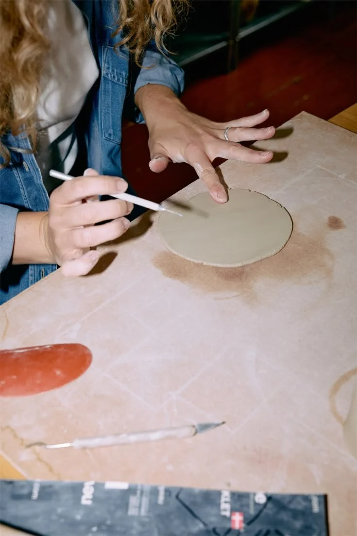 A person is working on a craft project, using a tool to paint or decorate a circular piece of material on a table. The person's hands are visible, with one hand holding the craft piece and the other using a tool.