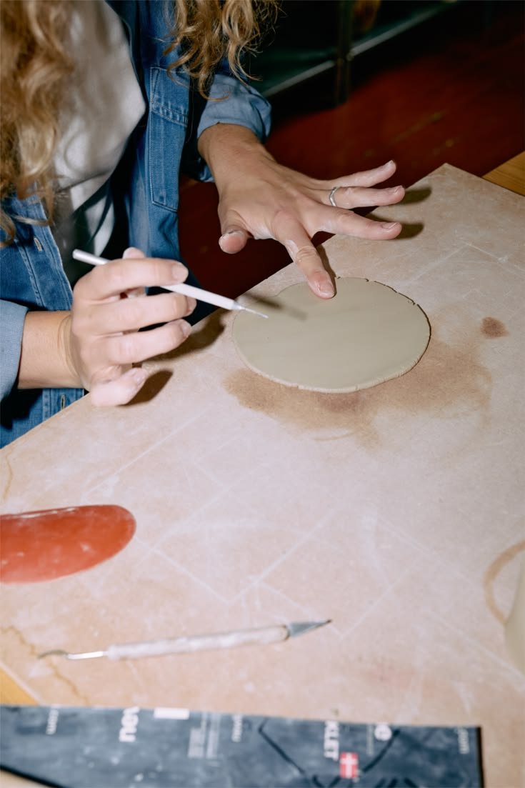 Person working on crafting or pottery with a round clay or ceramic piece on a table, using a small tool.