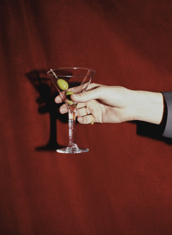 A hand holding a martini glass with a green olive on a toothpick against a red background.