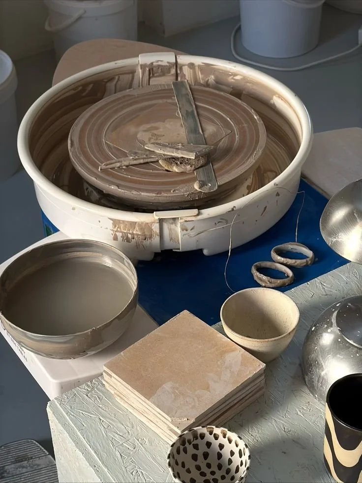 A pottery wheel with clay and tools, surrounded by bowls, tiles, and other ceramic items.