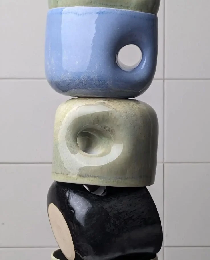 Stacked ceramic cups in blue, cream, and black, each with a circular indentation, set against a white tiled background.