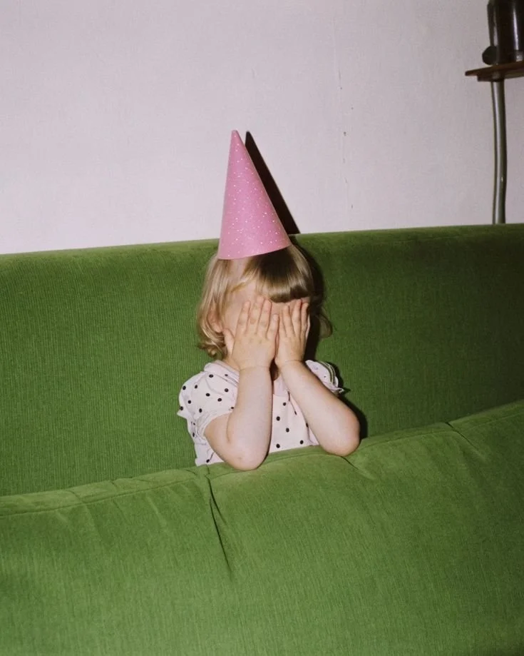 A young girl with blonde hair wearing a pink party hat and a polka dot shirt, covering her face with her hands while sitting on a green couch.