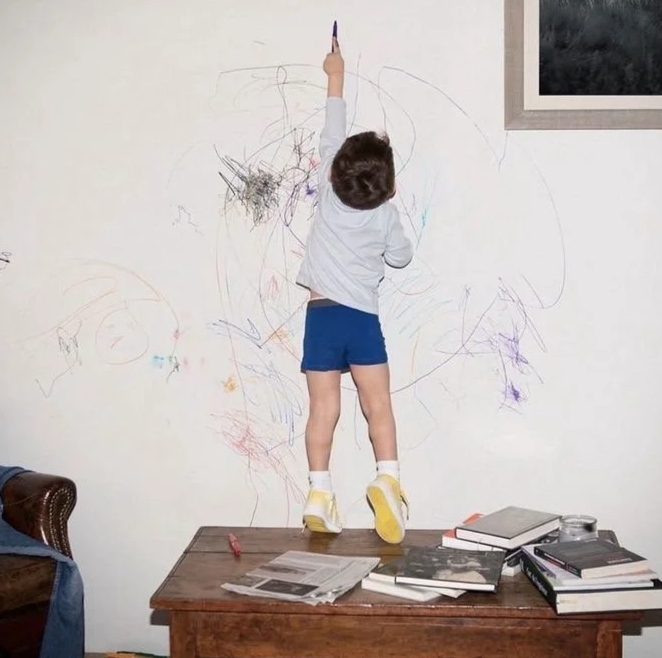 Child in blue shorts and yellow sneakers, drawing with a marker on a wall covered in colorful scribbles, standing on a wooden table with books and papers.