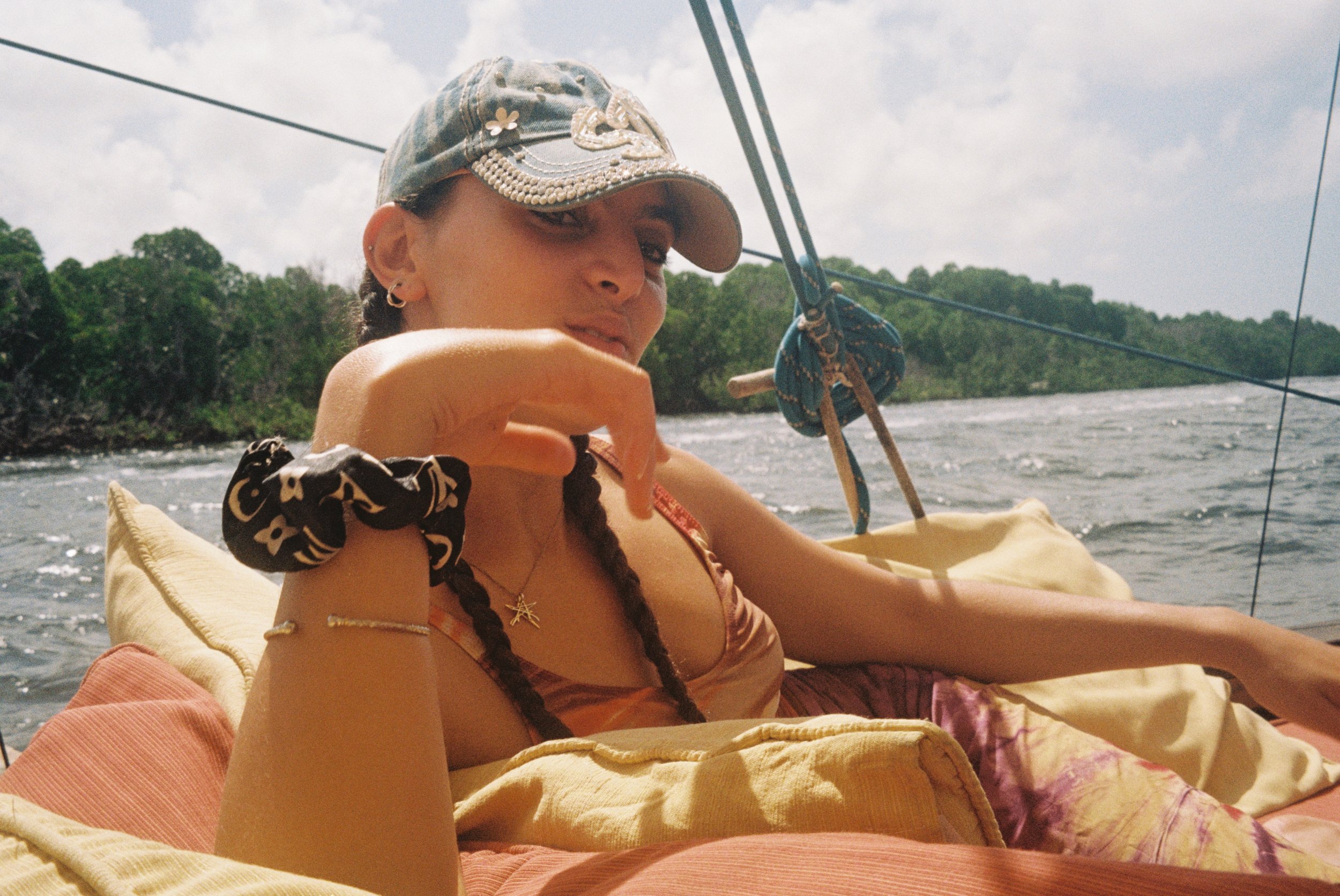 A woman with braided hair wearing a baseball cap, sitting on a boat, surrounded by cushions, with water and greenery in the background.