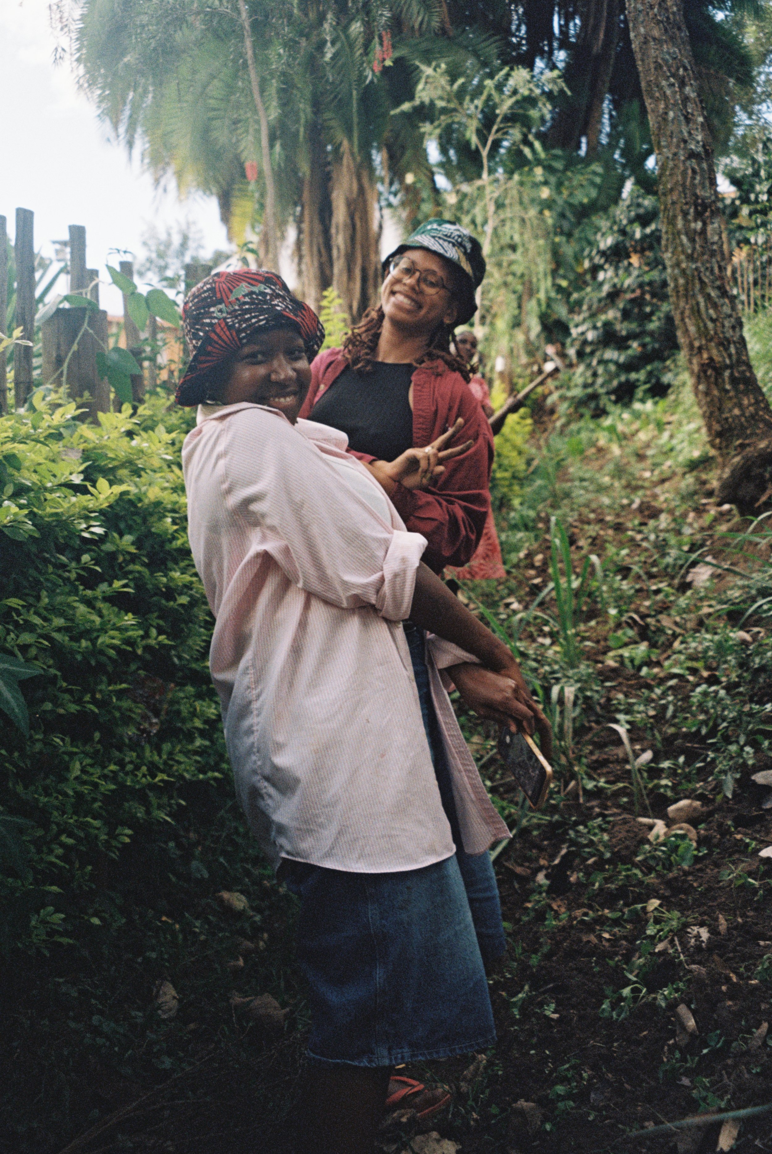 Two smiling women standing outdoors in a lush, green environment with trees and bushes, one holding a smartphone, both dressed casually and making peace signs.