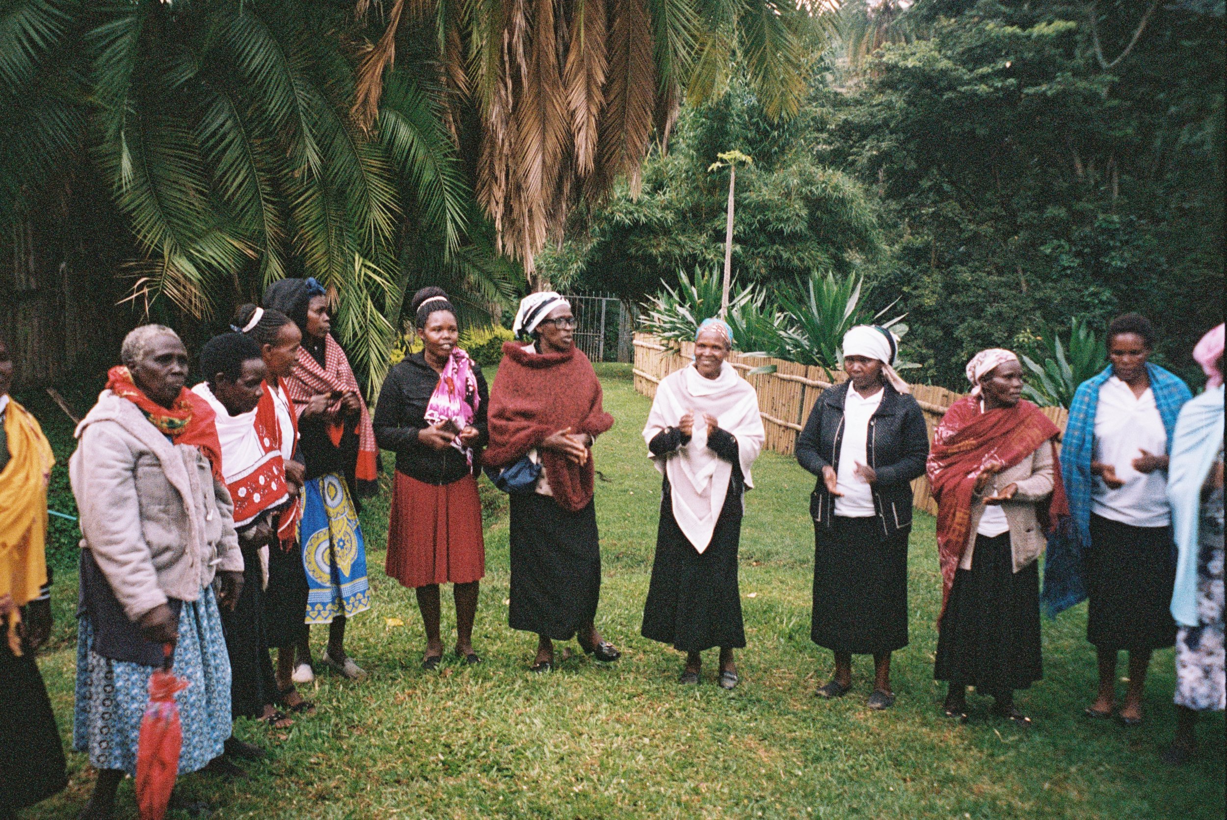 A group of women are standing in a circle outdoors in a lush green garden, engaged in conversation or a community activity.