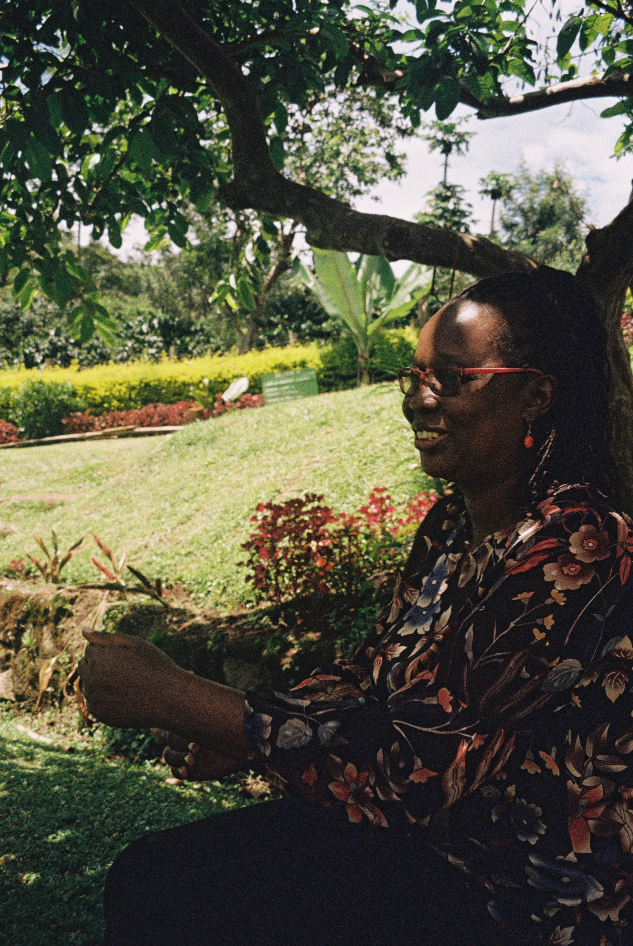 A woman with glasses and earrings sitting under a tree in a lush garden, smiling and holding a smartphone.
