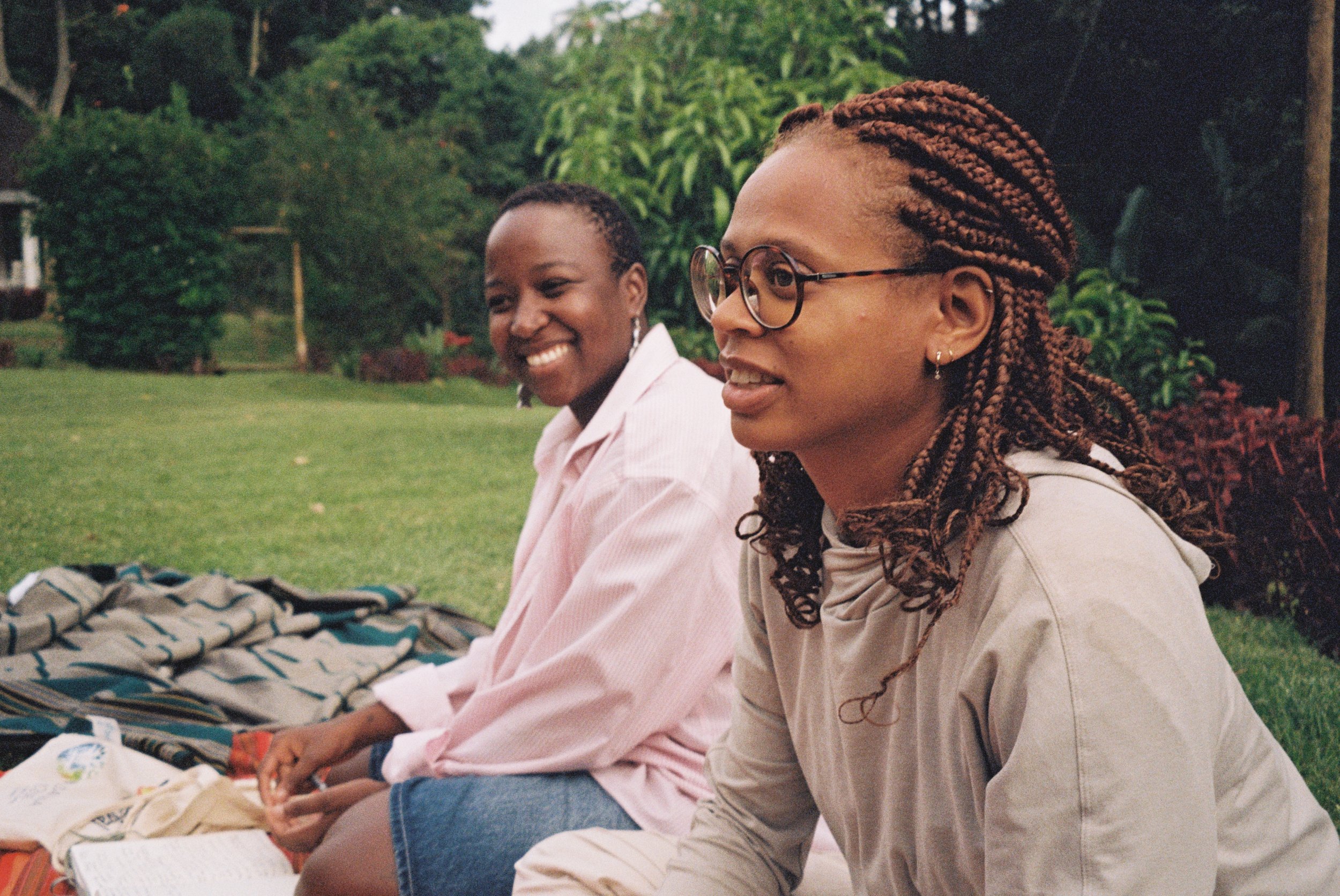Two women sitting on a blanket outdoors, smiling and conversing, with trees and plants in the background.