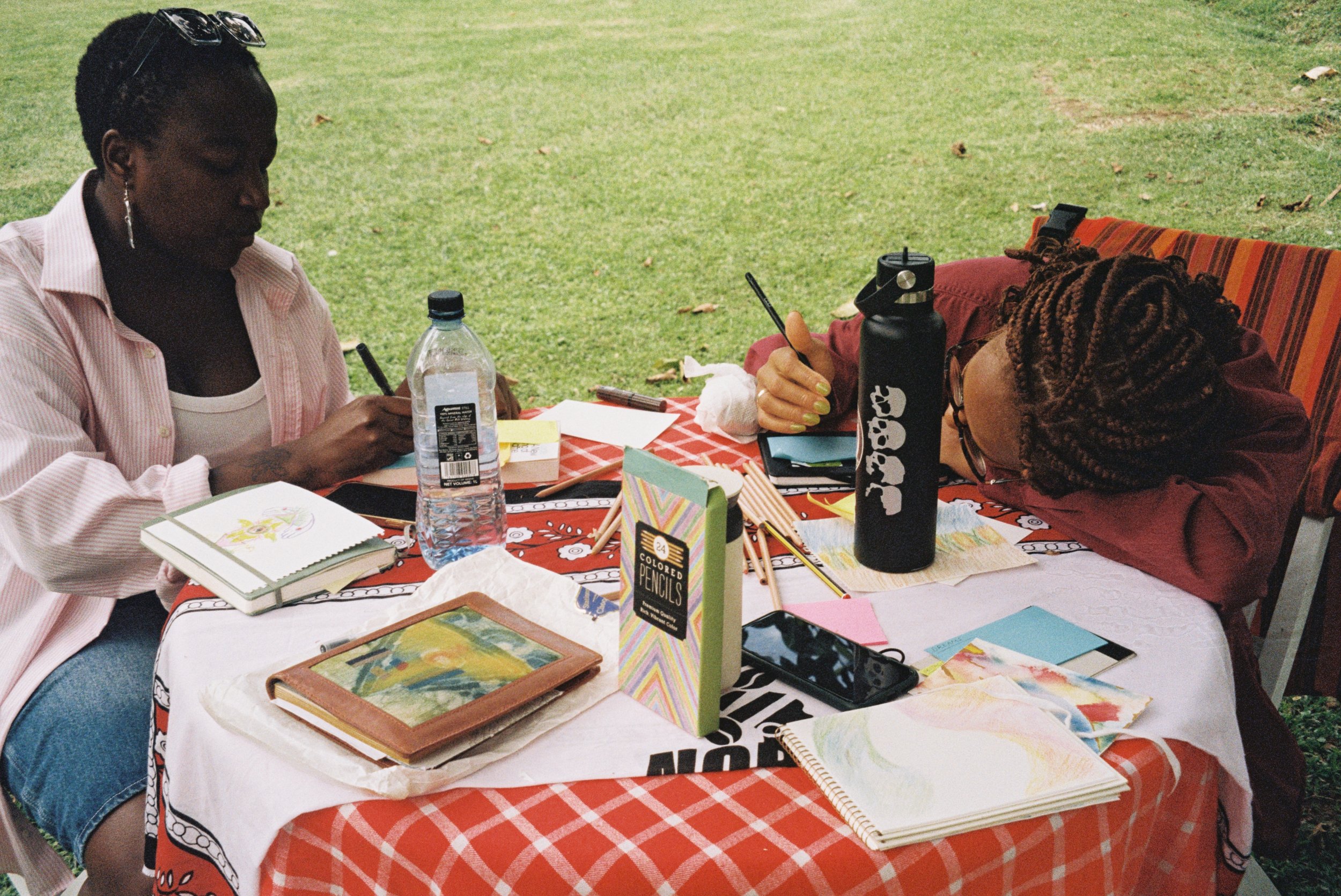 Two women sit at a table outdoors, drawing and coloring with art supplies such as colored pencils, notebooks, and sketchbooks, on a checked tablecloth with a grassy background.