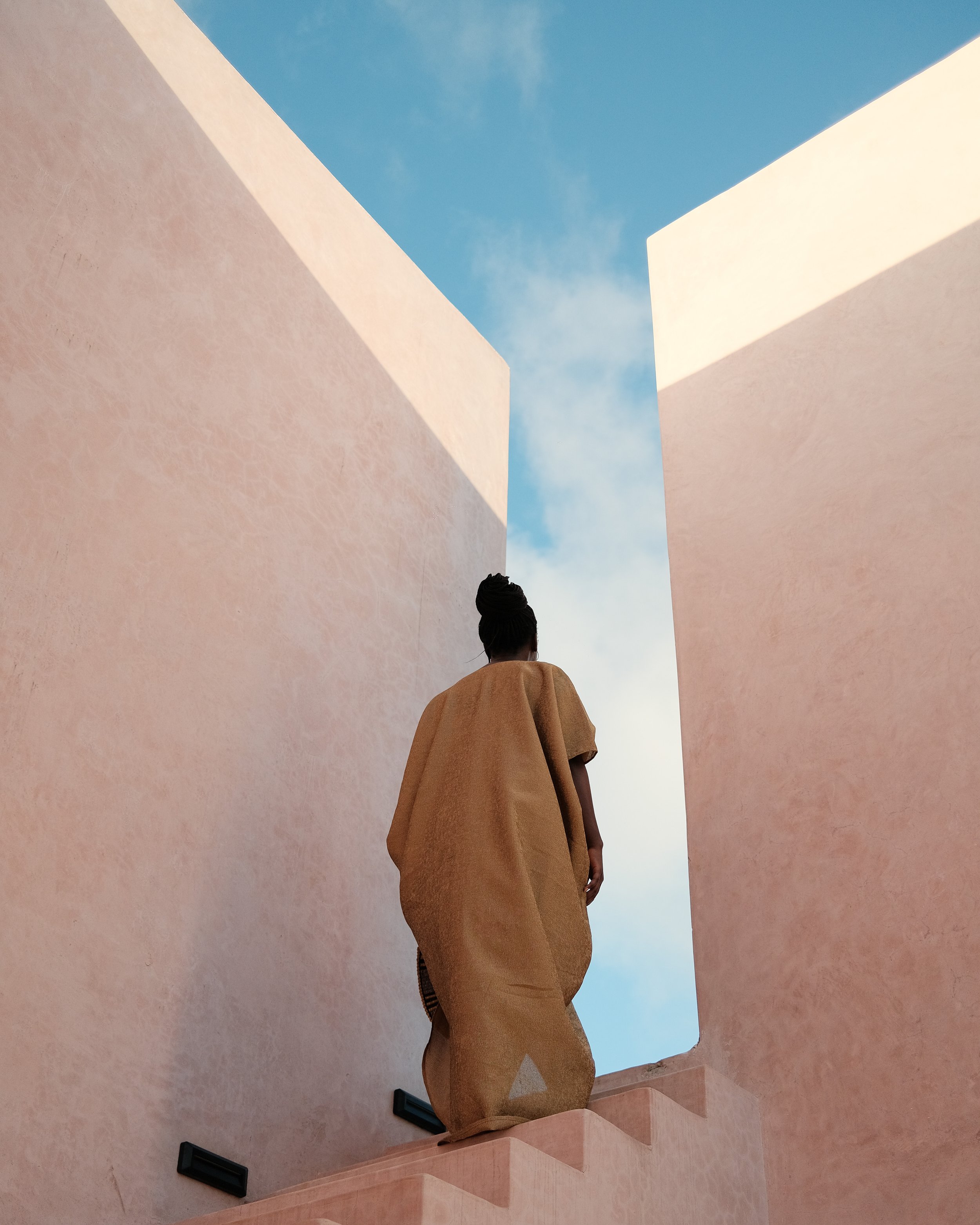 A person dressed in a brown garment walking up pink stairs between two pink walls, with a blue sky and clouds visible above.