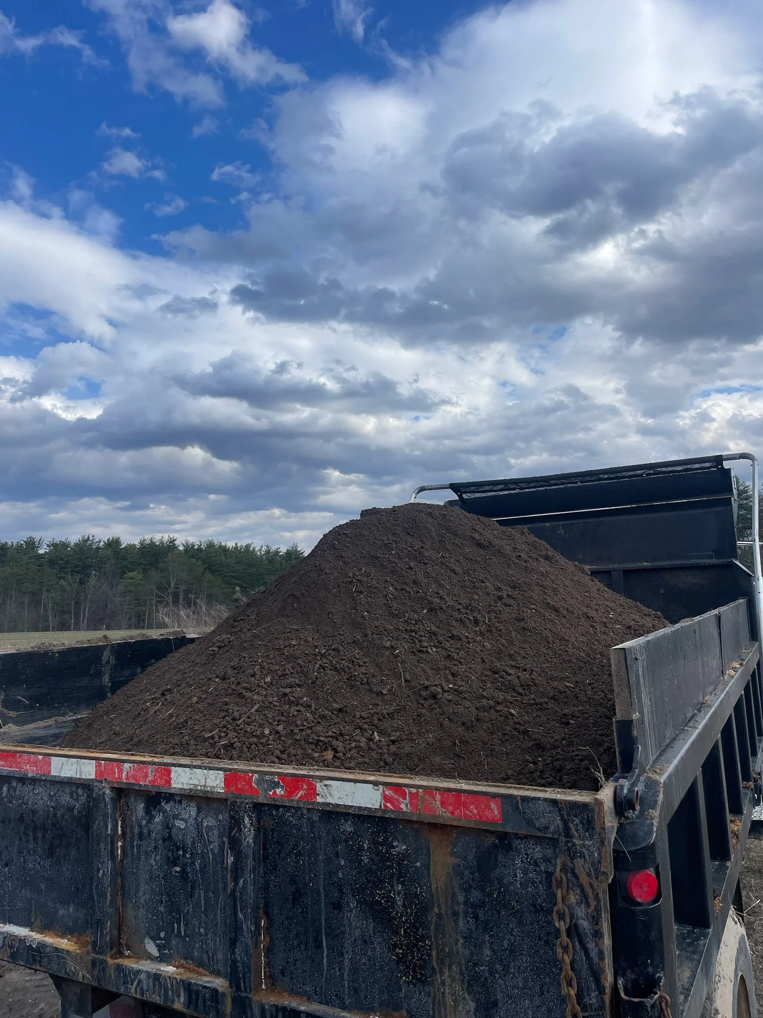 A truck loaded with fresh black soil or dirt on an outdoor construction or landscaping site, with a partly cloudy sky and a distant treeline in the background.