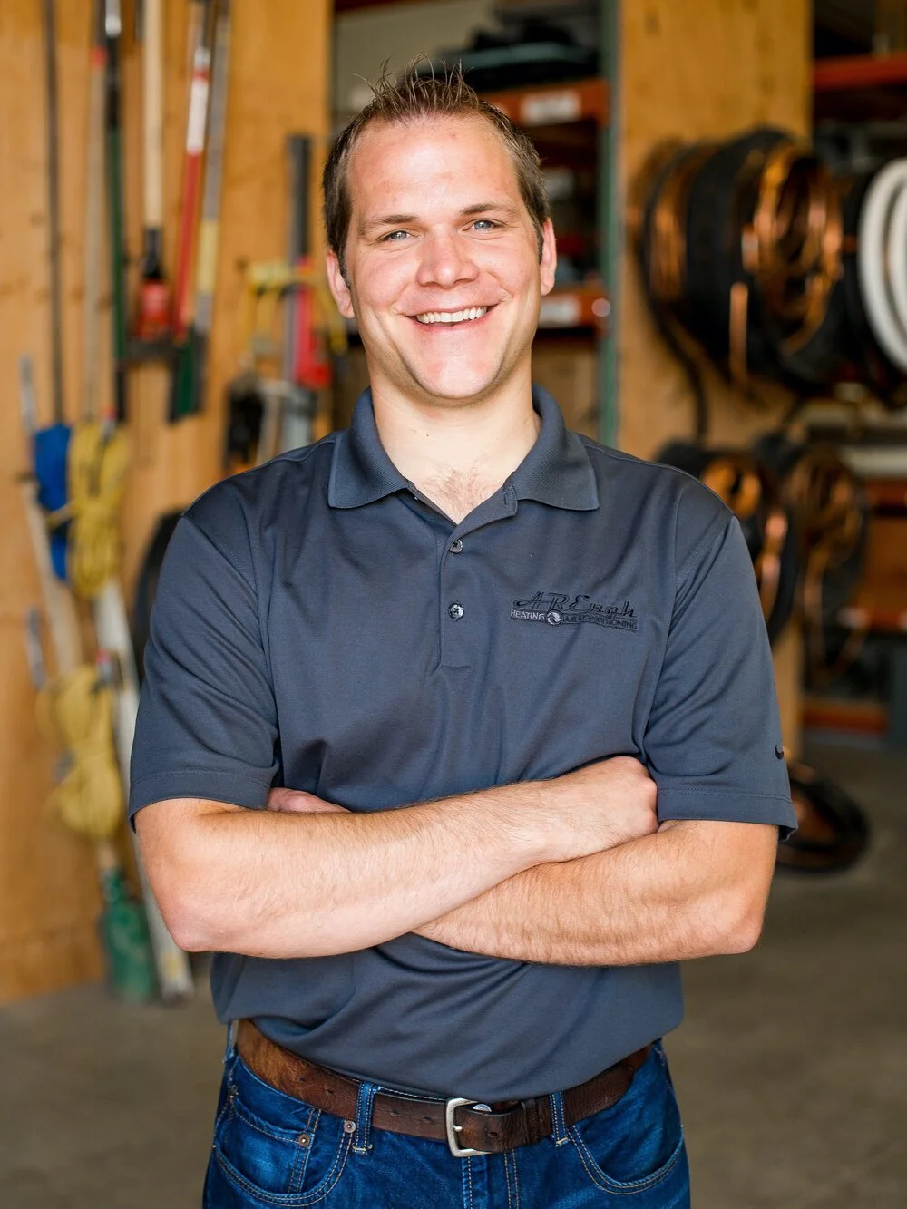 A smiling man with short brown hair standing in a workshop or garage with tools hanging on the wall behind him.