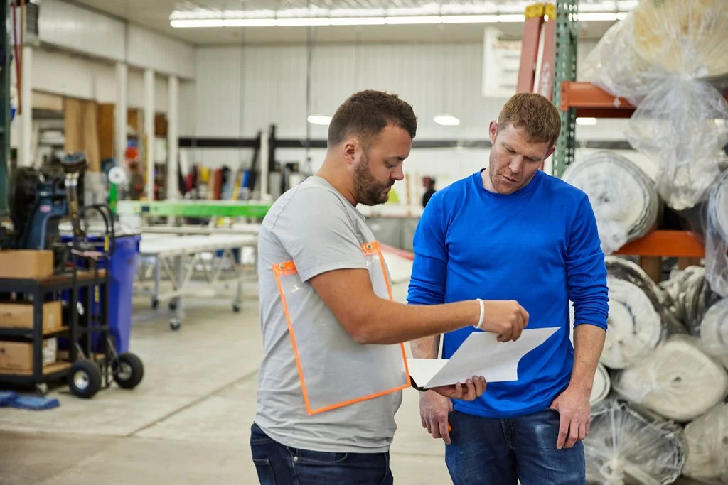 Two men in a warehouse or industrial space looking at a sheet of paper together.