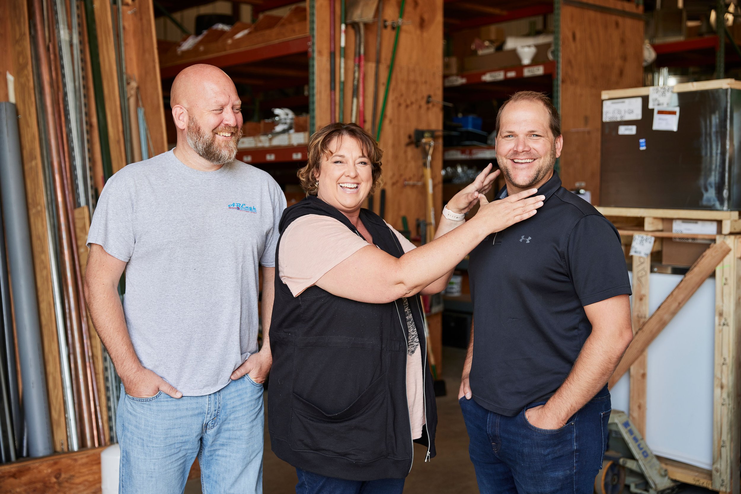 Three people standing inside a woodworking or hardware store, smiling and laughing. Two men and one woman, with shelves of tools and supplies in the background.
