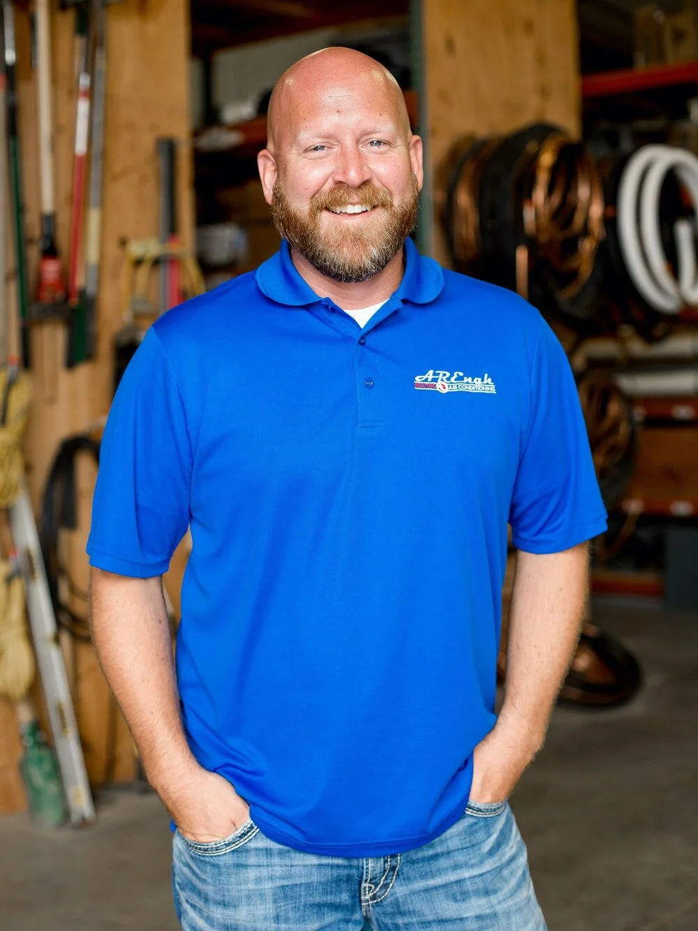 A smiling man with a beard wearing a blue polo shirt and jeans, standing in a workshop with tools and equipment in the background.