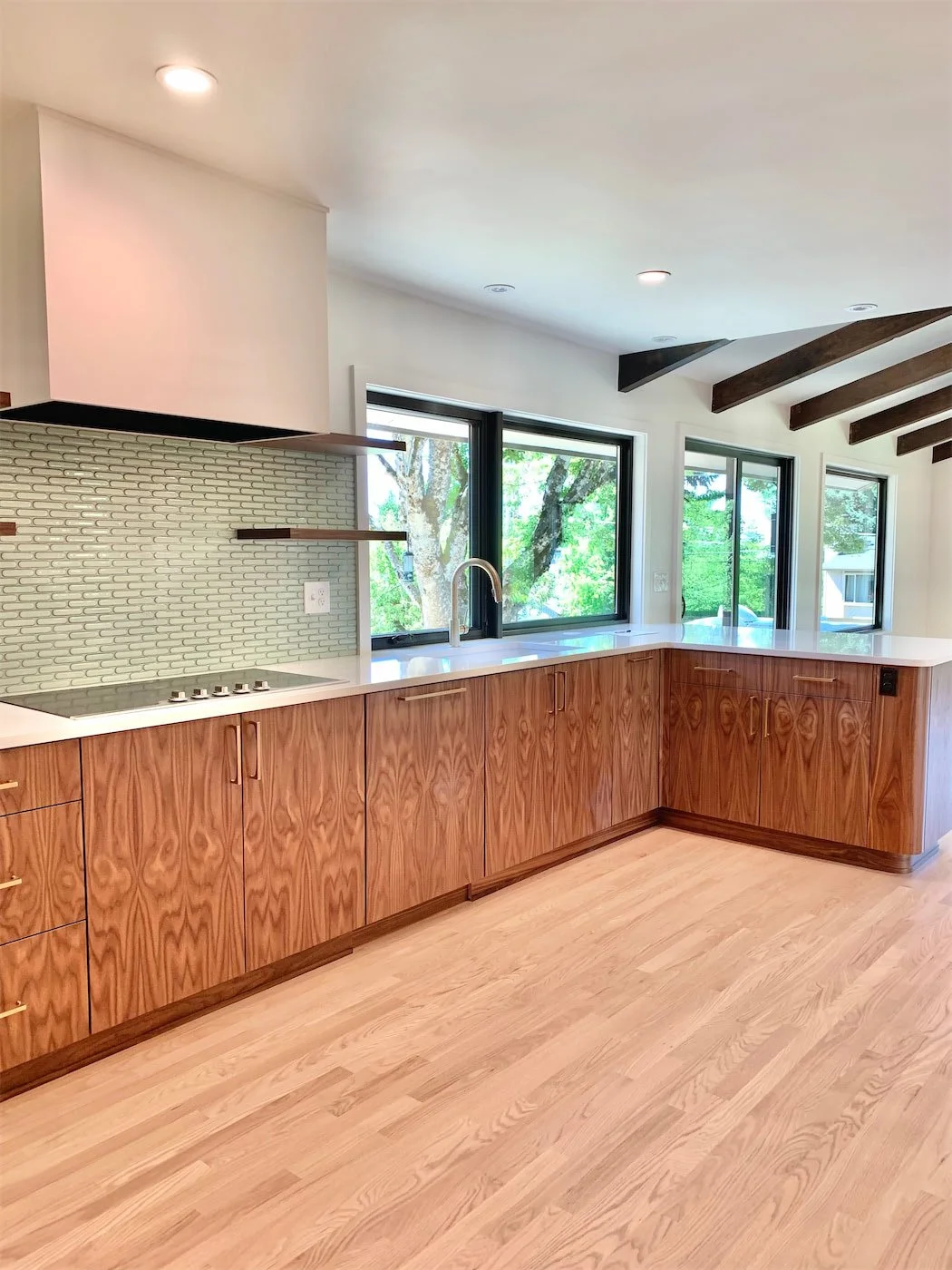 Modern kitchen with wooden cabinets, white countertops, a green tile backsplash, and large windows overlooking trees.