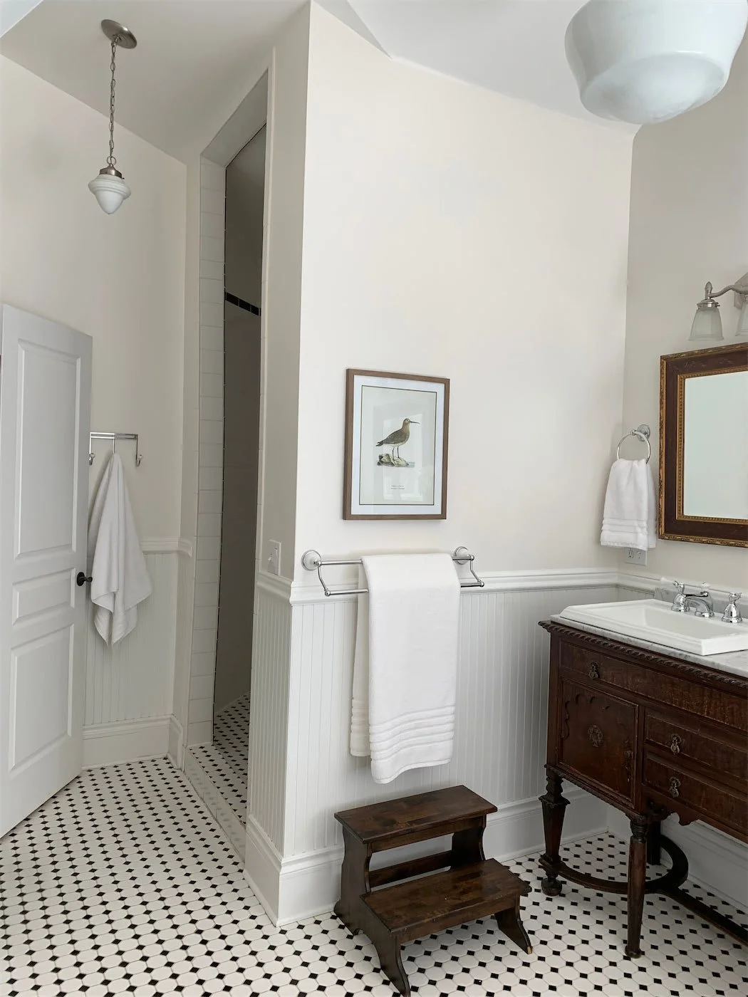 A bathroom with white walls and black-and-white hexagonal tiled floor. It features a wooden vanity with a white sink, a framed bird illustration on the wall, and a grate on the wall with white towels hanging. There is a small dark wood step ladder, and lighting fixtures, including a pendant lamp and wall sconces.