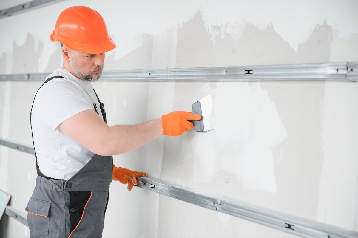 A man in a construction worker outfit, wearing an orange safety helmet and orange gloves, smoothing drywall with a hand trowel.