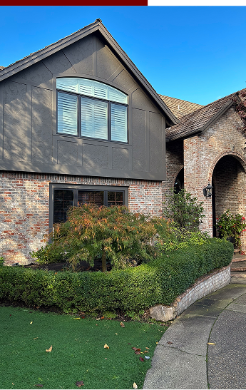 Close-up of a suburban house with brick and dark gray siding, large front window, well-maintained front yard with bushes, small tree, and a curved pathway, under a clear blue sky.