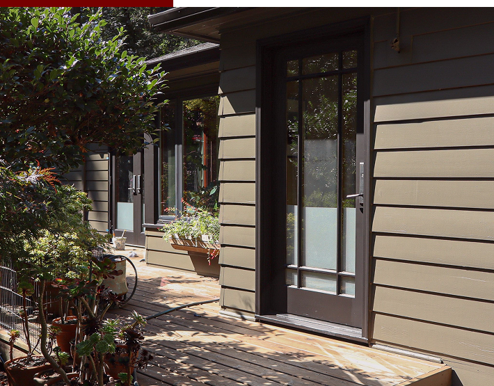 Exterior view of a house with beige siding, large glass door, and a wooden deck. There are potted plants and greenery on the deck.