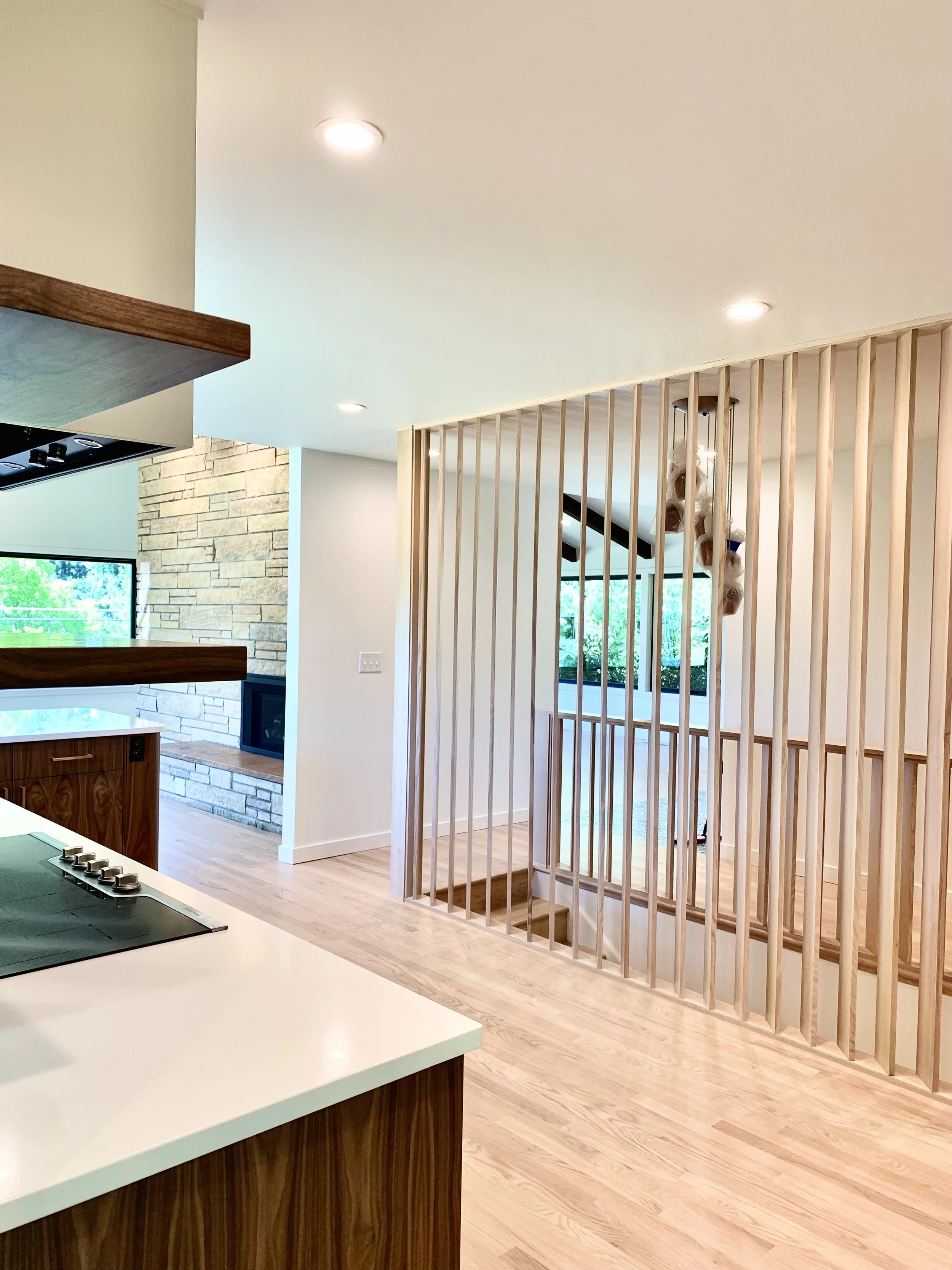 Interior view of a modern house with a wooden staircase enclosed by vertical wooden slats, a kitchen with a white countertop, and a brick fireplace in the background.