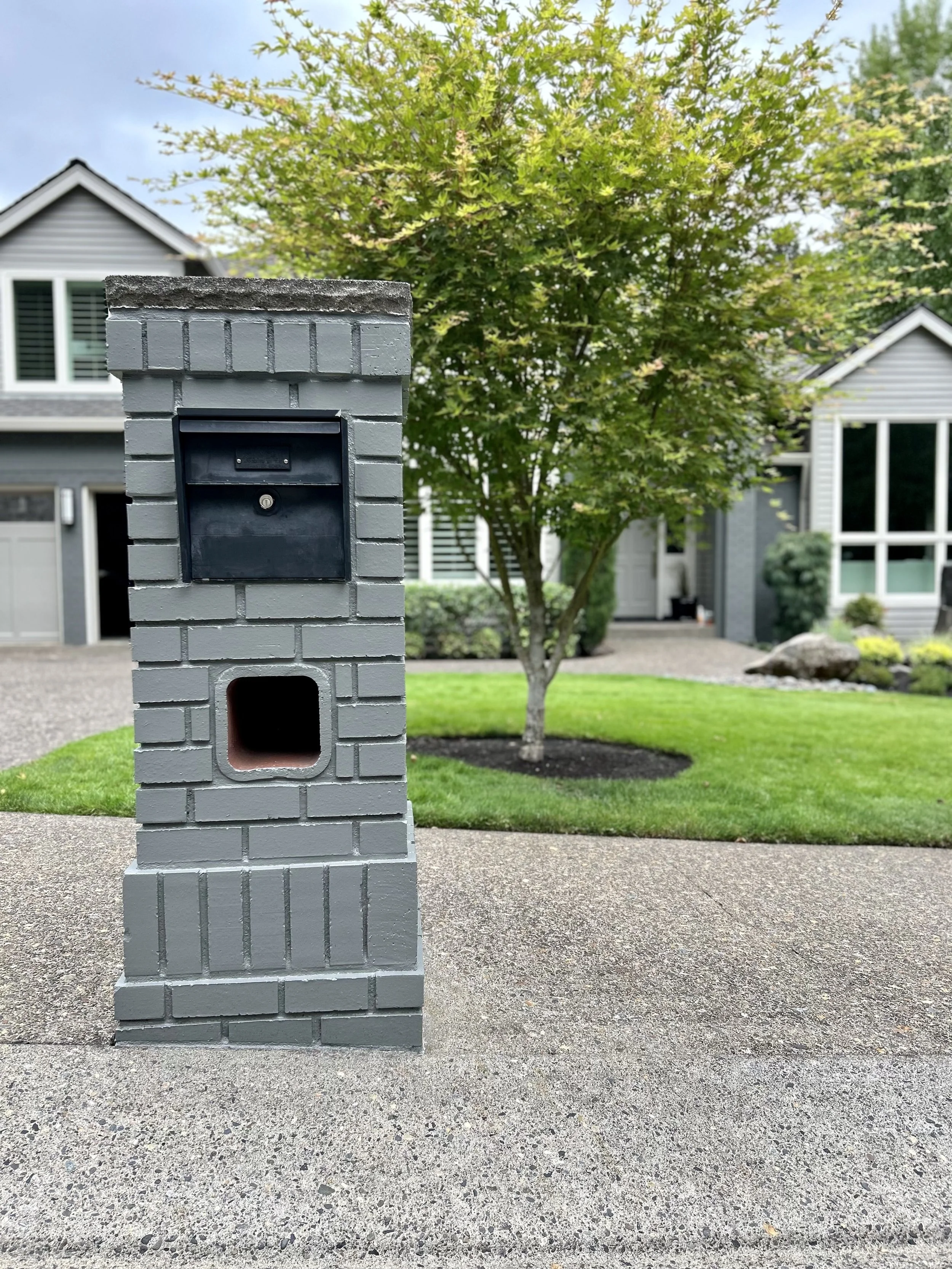 Gray brick mailbox post with a black mailbox on top and a small open compartment below, set on a concrete sidewalk in front of a landscaped yard with a green lawn, a tree, and houses in the background.