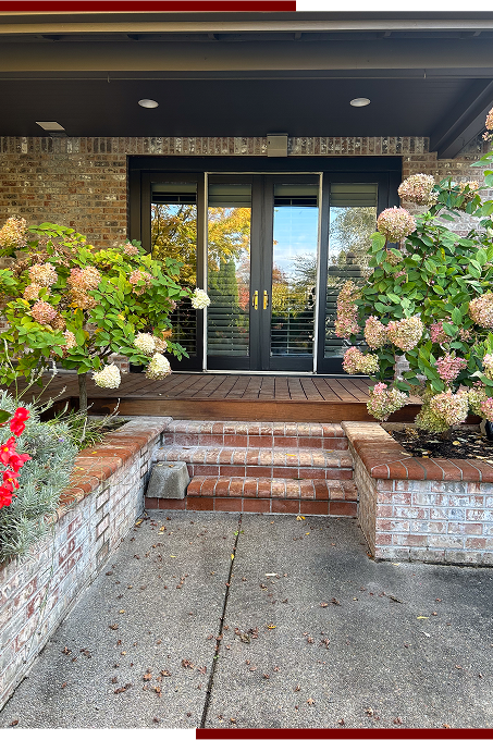 Front porch with brick stairs leading to glass double doors, surrounded by large flowering bushes, concrete sidewalk, and brick house exterior.