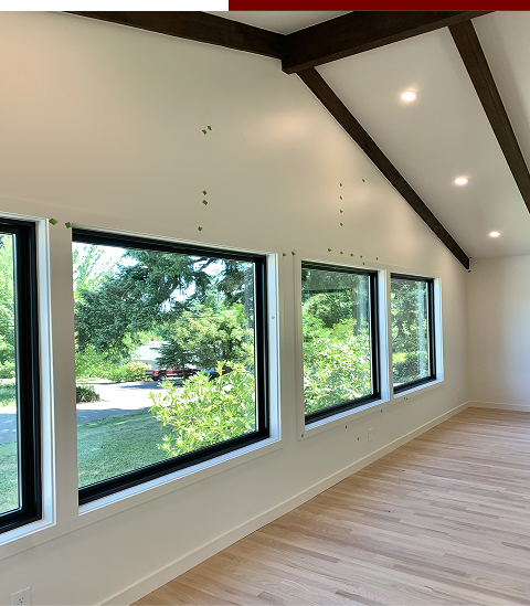 Empty room with three large windows looking outside at greenery, white walls, light wooden floor, and dark wooden beams on the ceiling.