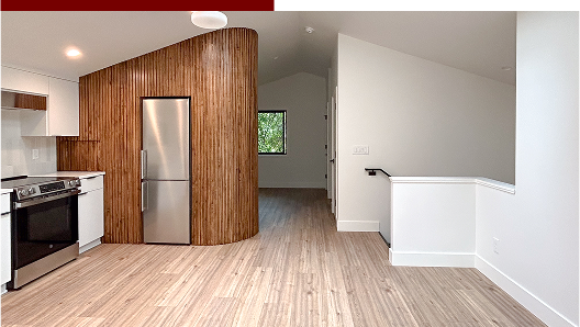 Interior view of a modern kitchen with wooden accents, stainless steel refrigerator, and light wood flooring, adjacent to a white wall with a staircase.