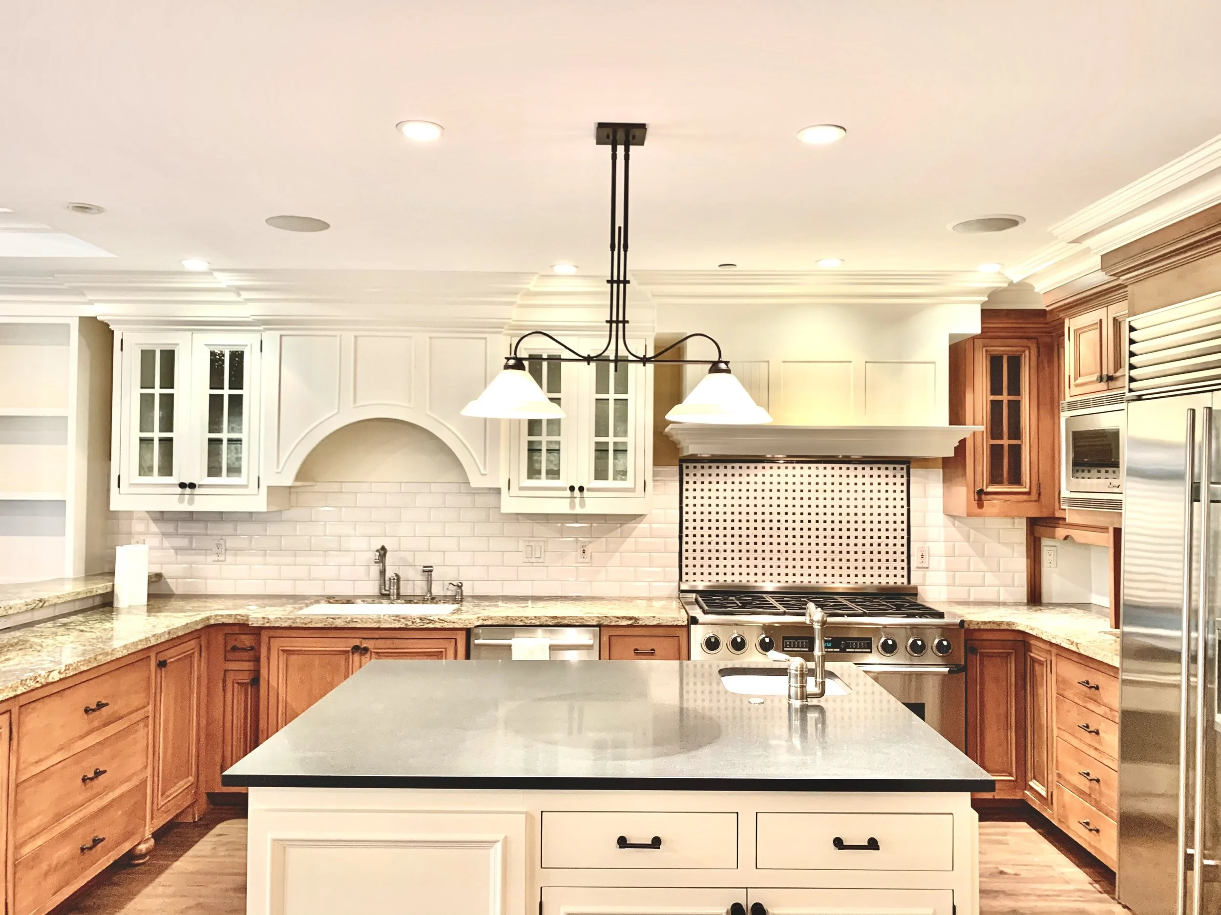 Modern kitchen with white and wooden cabinetry, granite countertops, stainless steel appliances, a kitchen island with a sink, and pendant lighting.