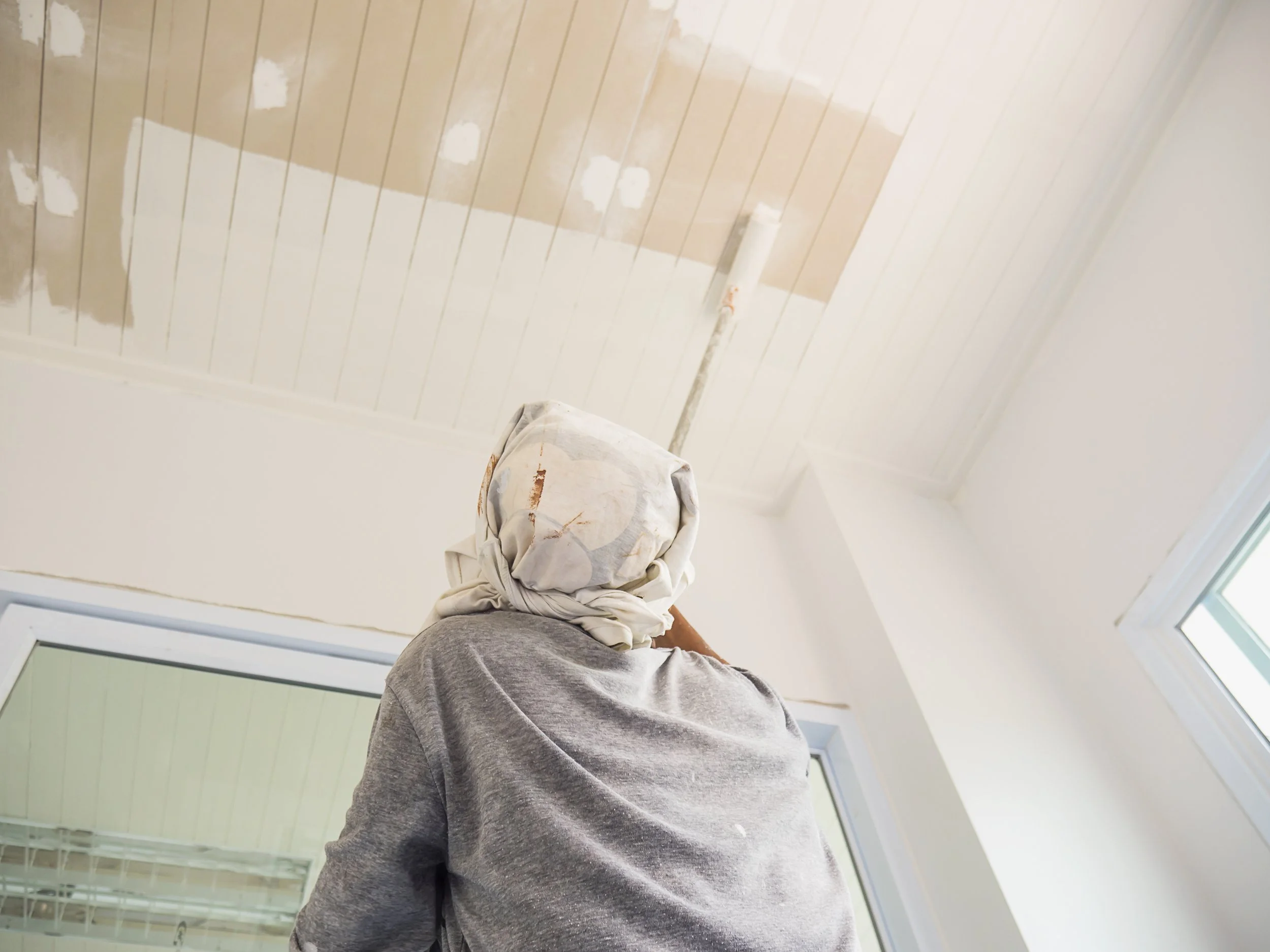 Person painting a ceiling with wood paneling, wearing a headscarf and gray shirt, using a roller brush near a window.