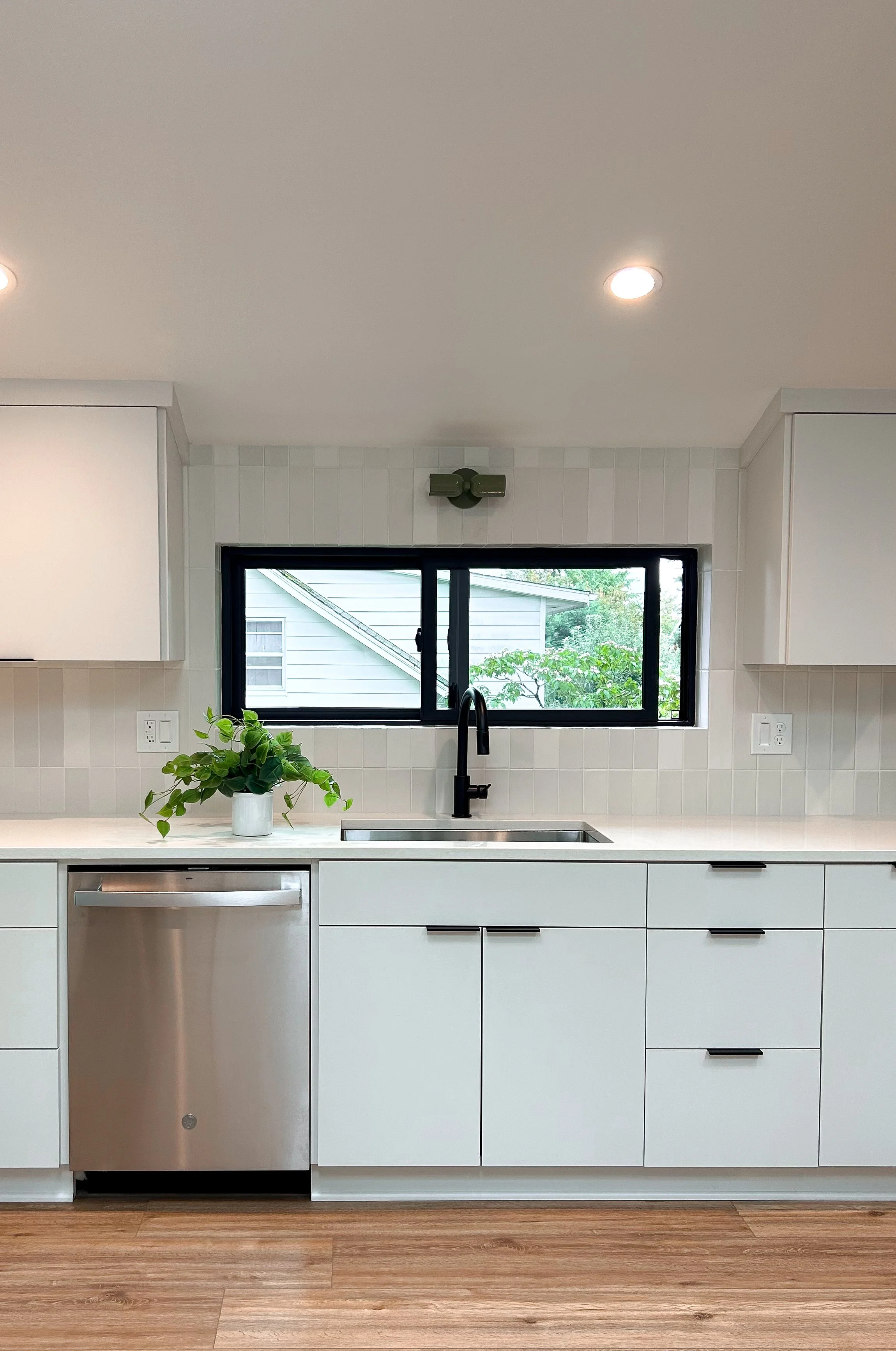 Modern kitchen with white cabinetry, a black faucet, and a black window overlooking greenery and a neighboring house, with a potted plant on the counter.
