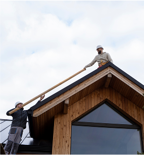 Two workers on a wooden house roof, one standing on the roof and the other on the side, holding a long pole.