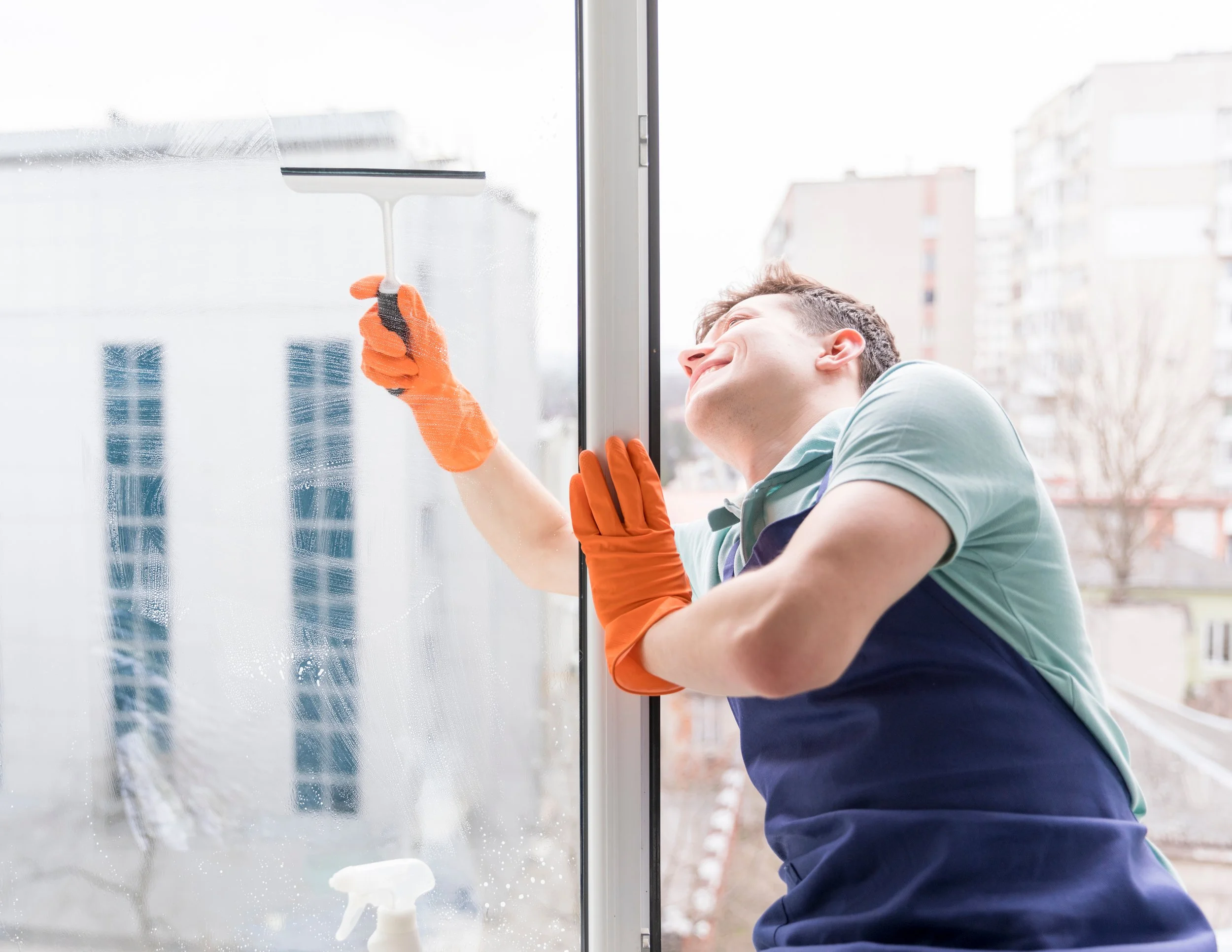 A man cleaning a window with a squeegee inside a building, wearing an apron and orange gloves, with a cityscape in the background.