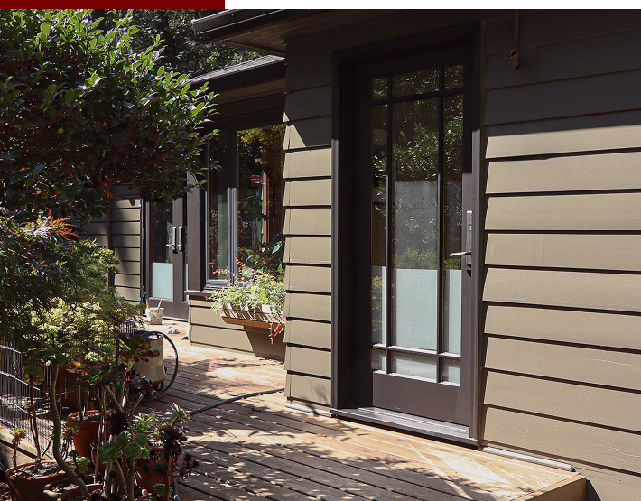 Exterior of a house with a wooden deck, sliding glass door, beige siding, surrounded by plants and trees.