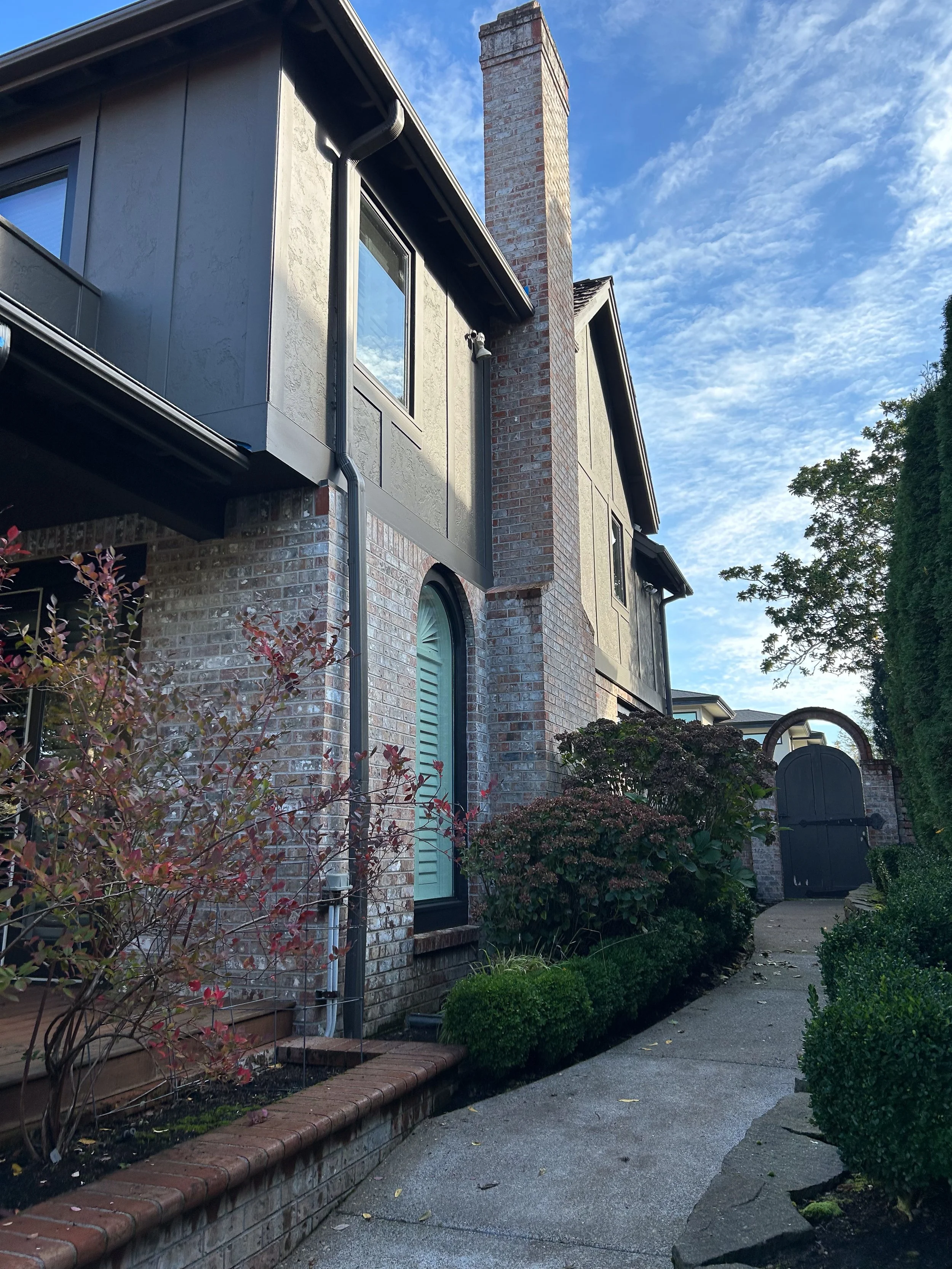 A view of a house with brick walls, a chimney, and a pathway leading to a gate, surrounded by plants and bushes.
