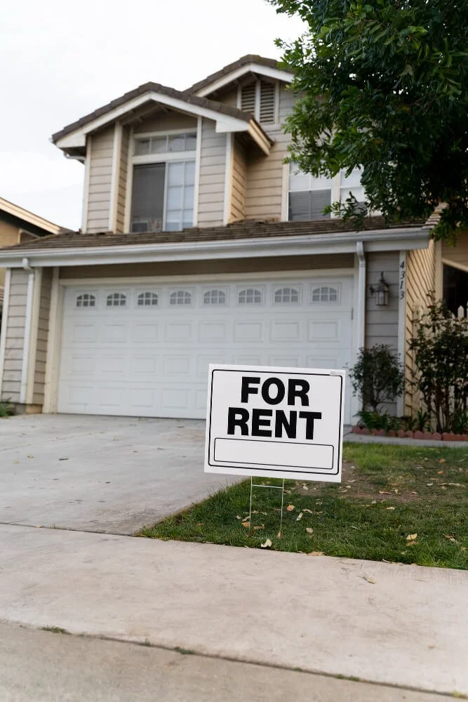 A two-story house with beige siding, white garage door, and a 'For Rent' sign in the front yard.