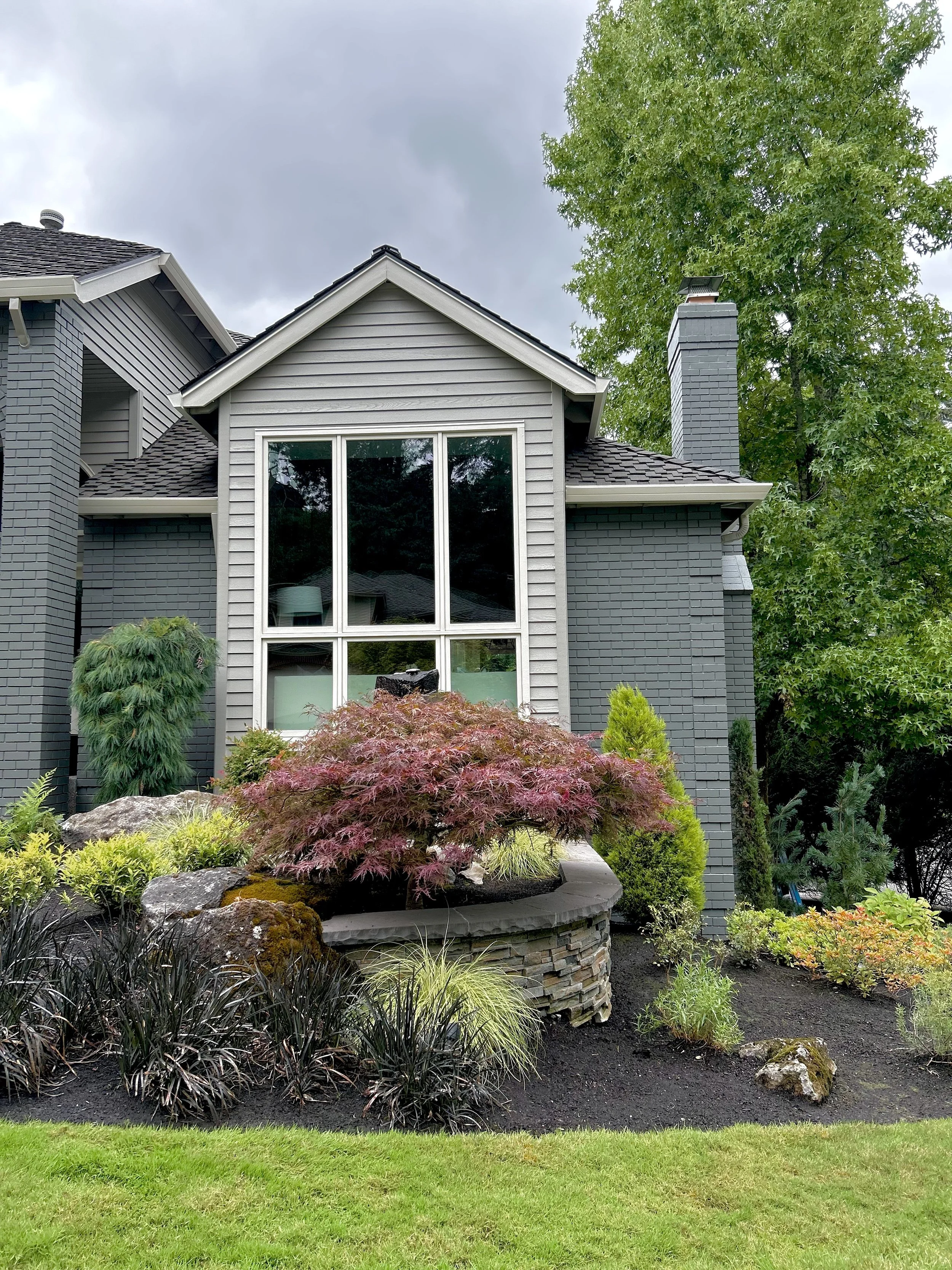 Front view of a gray house with a large window, surrounded by landscaped garden with a Japanese maple tree, rocks, and various shrubs, under an overcast sky.
