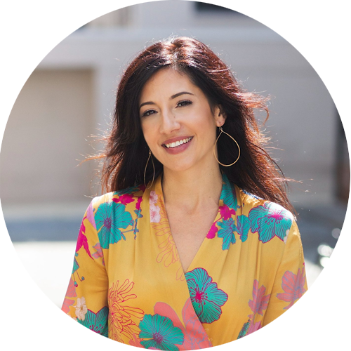 A woman with long dark hair smiling outdoors, wearing a yellow floral blouse and large hoop earrings.