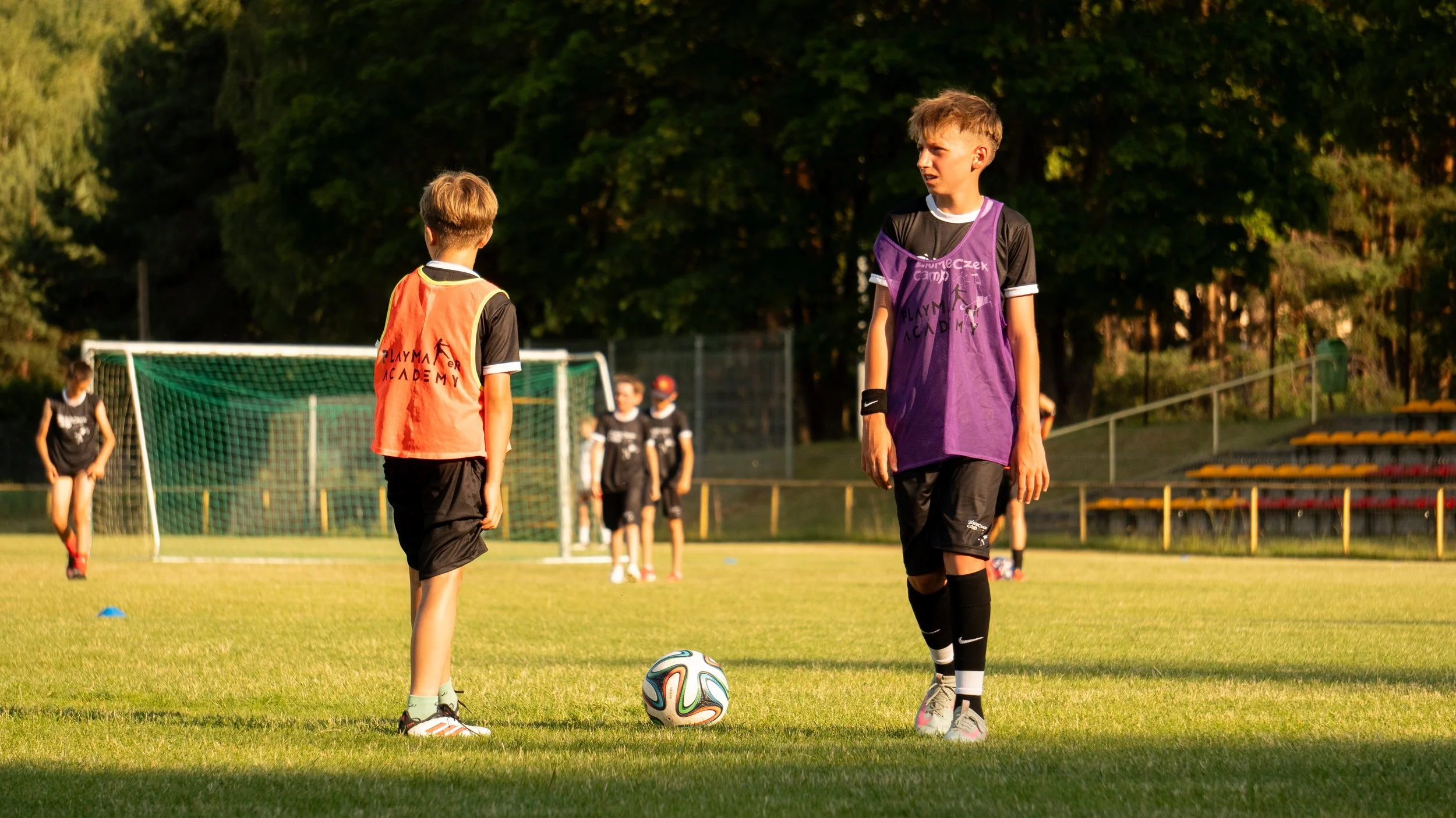 Dzieci grają w piłkę nożną na boisku. Jeden chłopiec przygotowuje się do kopnięcia piłki, a w tle widać innych zawodników w strojach sportowych. Scena rozgrywa się prawdopodobnie późnym popołudniem lub wczesnym wieczorem.