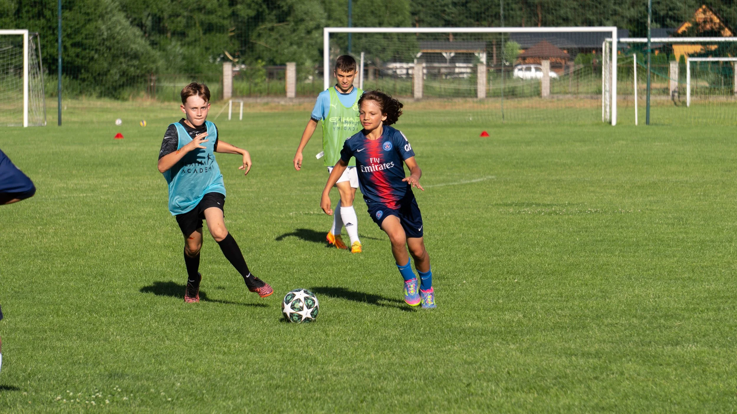 Dzieci grają w piłkę nożną na trawiastym boisku z bramkami w tle. Chłopak w koszulce Paris Saint-Germain biegnie za piłką, chłopiec w turkusowej kamizelce treningowej również ją goni, a trzecie dziecko w niebieskiej kamizelce obserwuje akcję z tyłu