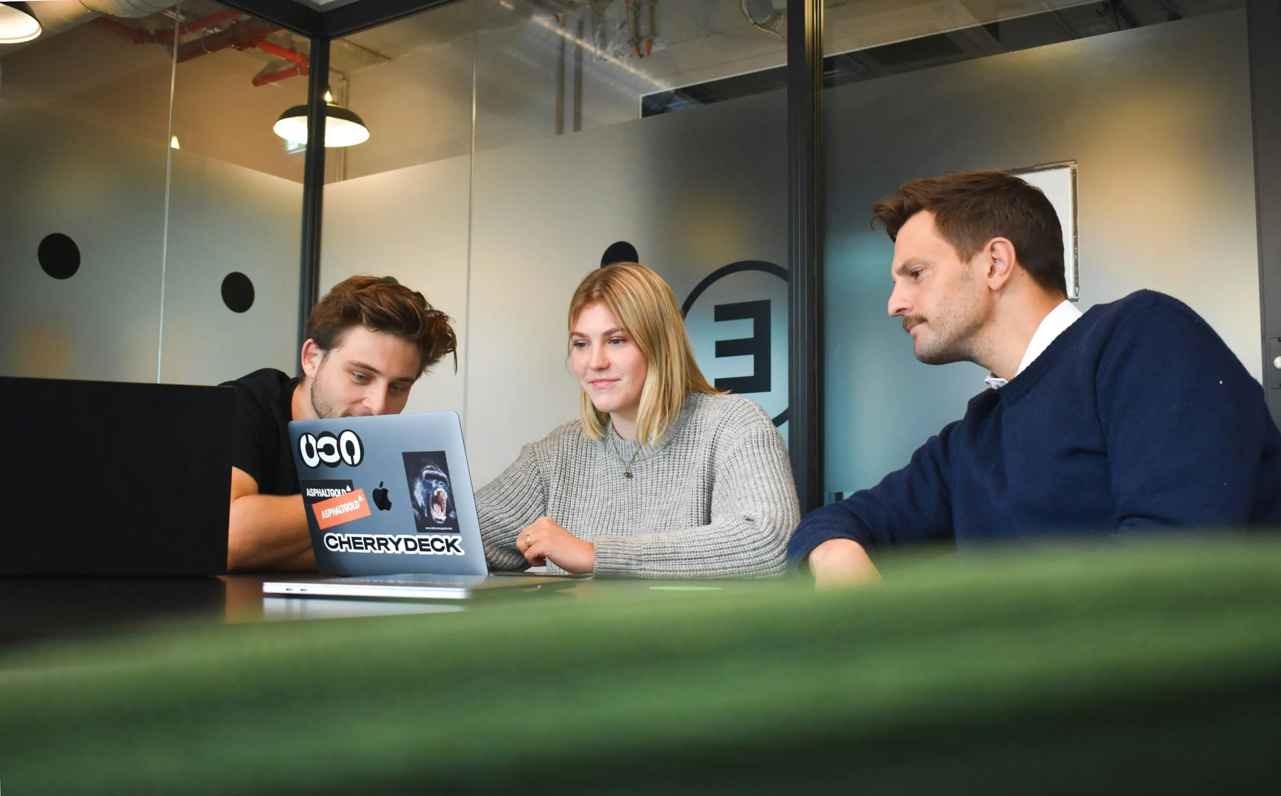 Three people sitting closely at a table, looking at a laptop screen, in an office or conference room setting. They are looking how to boost their careers.