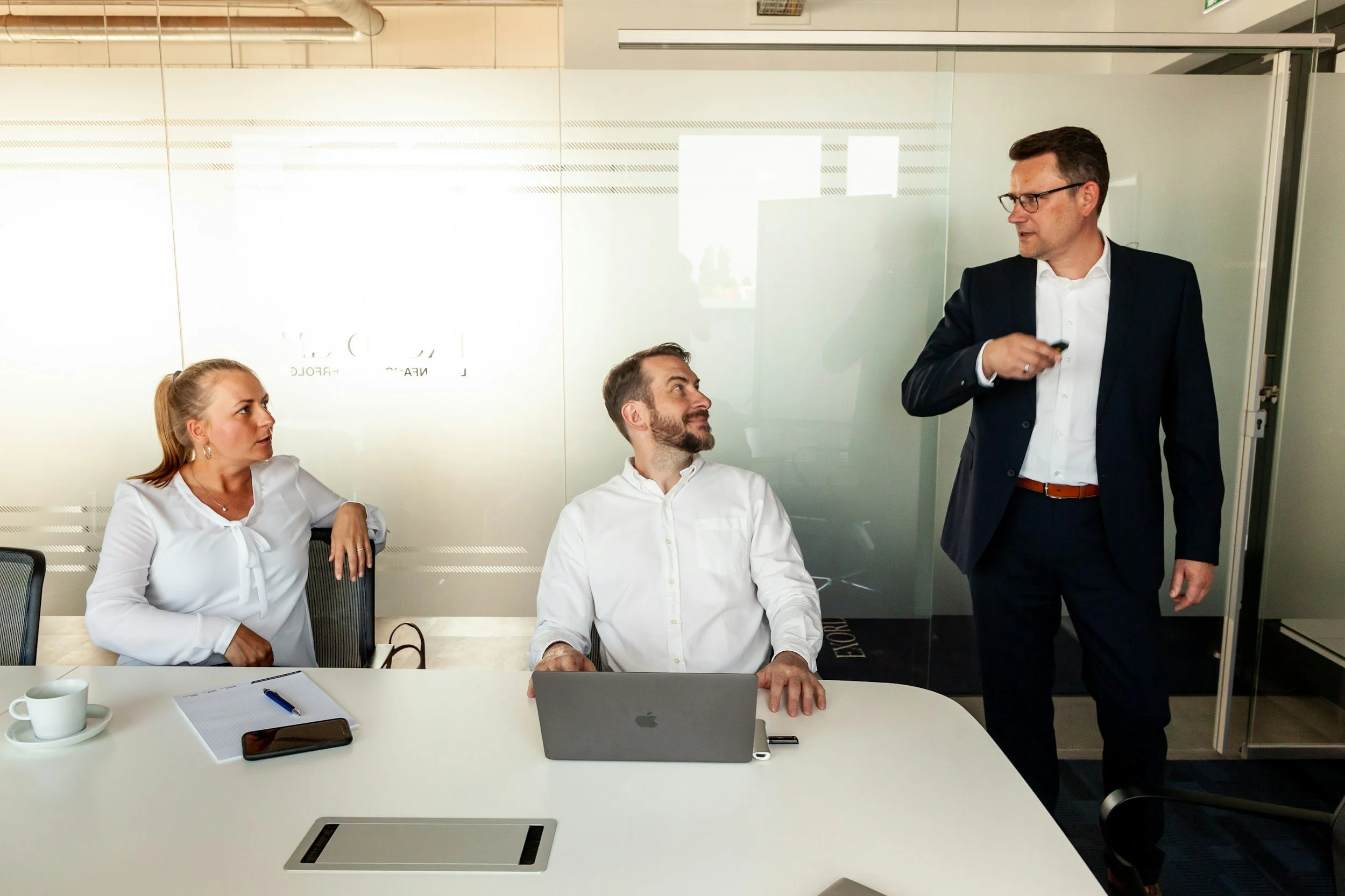 A professional office scene with three individuals having a discussion. The woman on the left is sitting at a conference table with a cup, a notebook, and a smartphone. The man in the middle, also seated, is looking up at the man standing. The standing man is wearing a dark suit and glasses, holding a presentation remote, and appears to be speaking to the group. There is a laptop on the table and a glass wall in the background.
