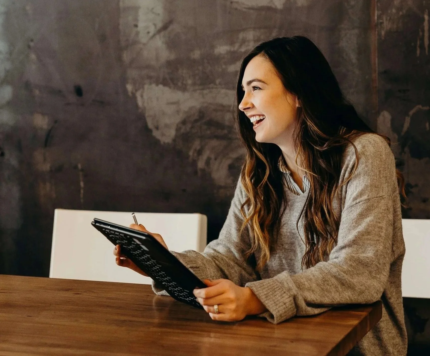 A woman client of Career Evolution sitting at a wooden table, smiling and holding a calculator in her hands. She has long, wavy dark hair, and is wearing a beige sweater. The background features a dark, textured wall.