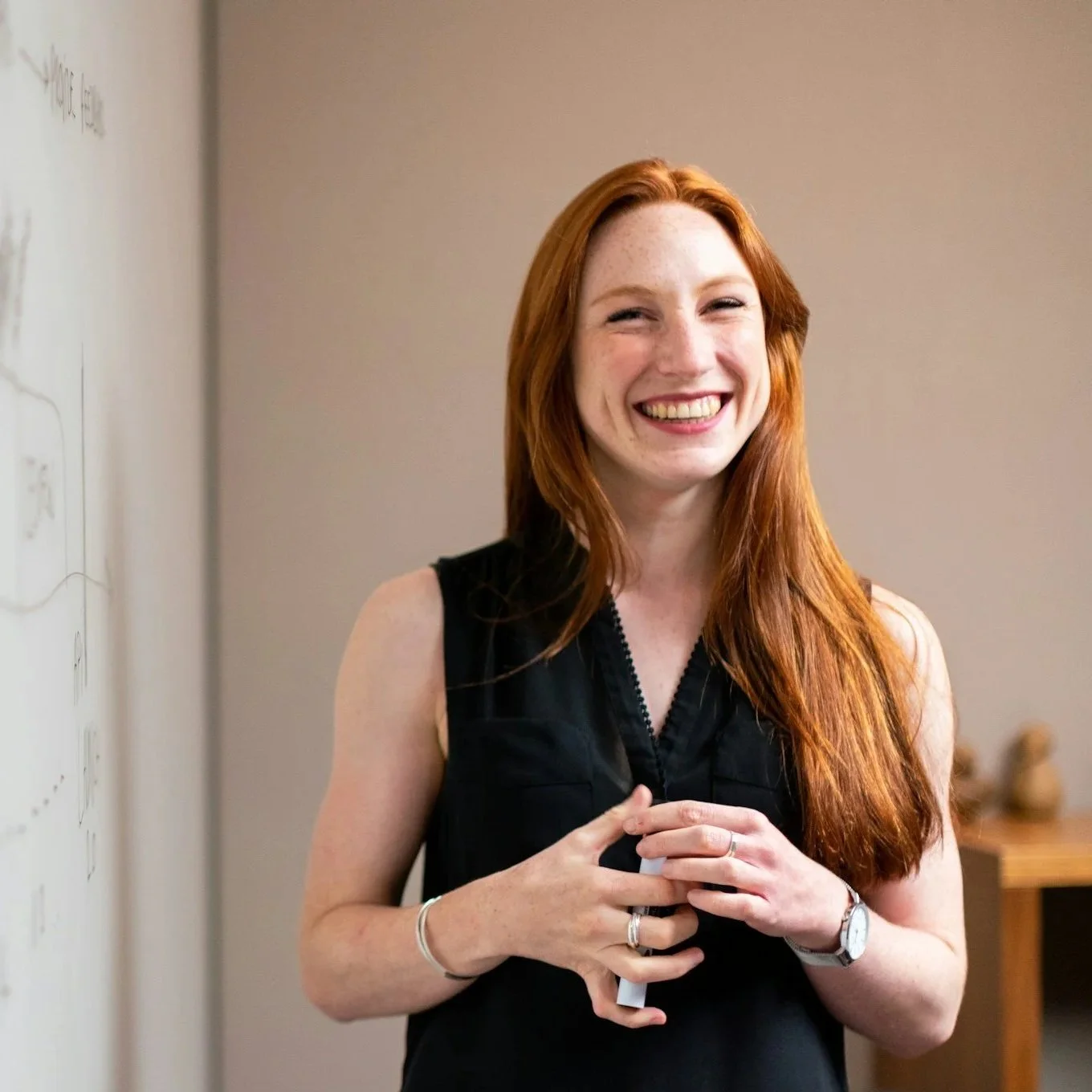A woman with long red hair and a black sleeveless top smiling and holding a white object, standing indoors near a whiteboard. she is excitedly looking for her next job move after talking to her career coach at Career Evolution.