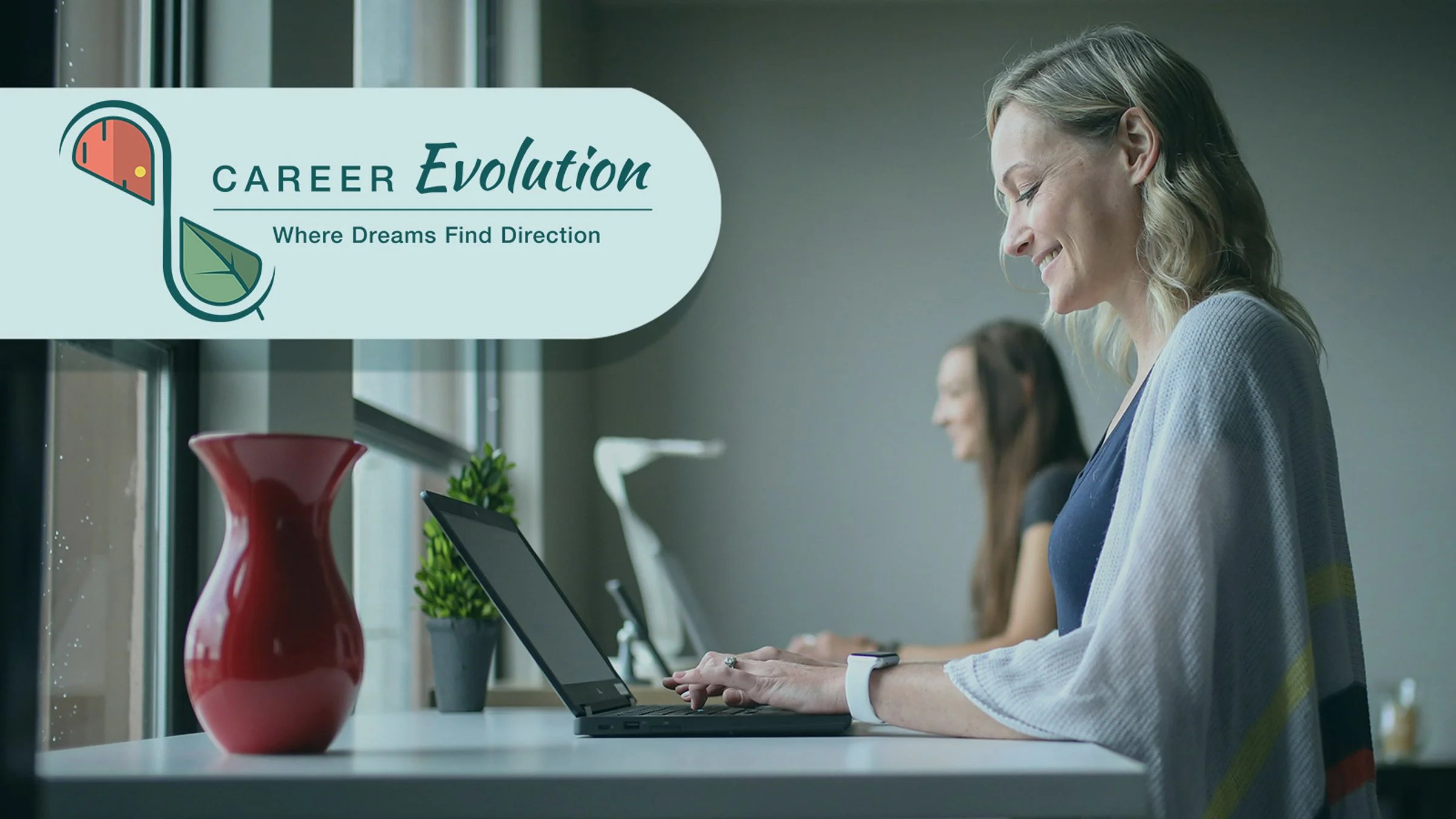 A woman coaching client of Career Evolution smiling while working on a laptop and working on her career moves at a desk with decorative items and a window in the background.