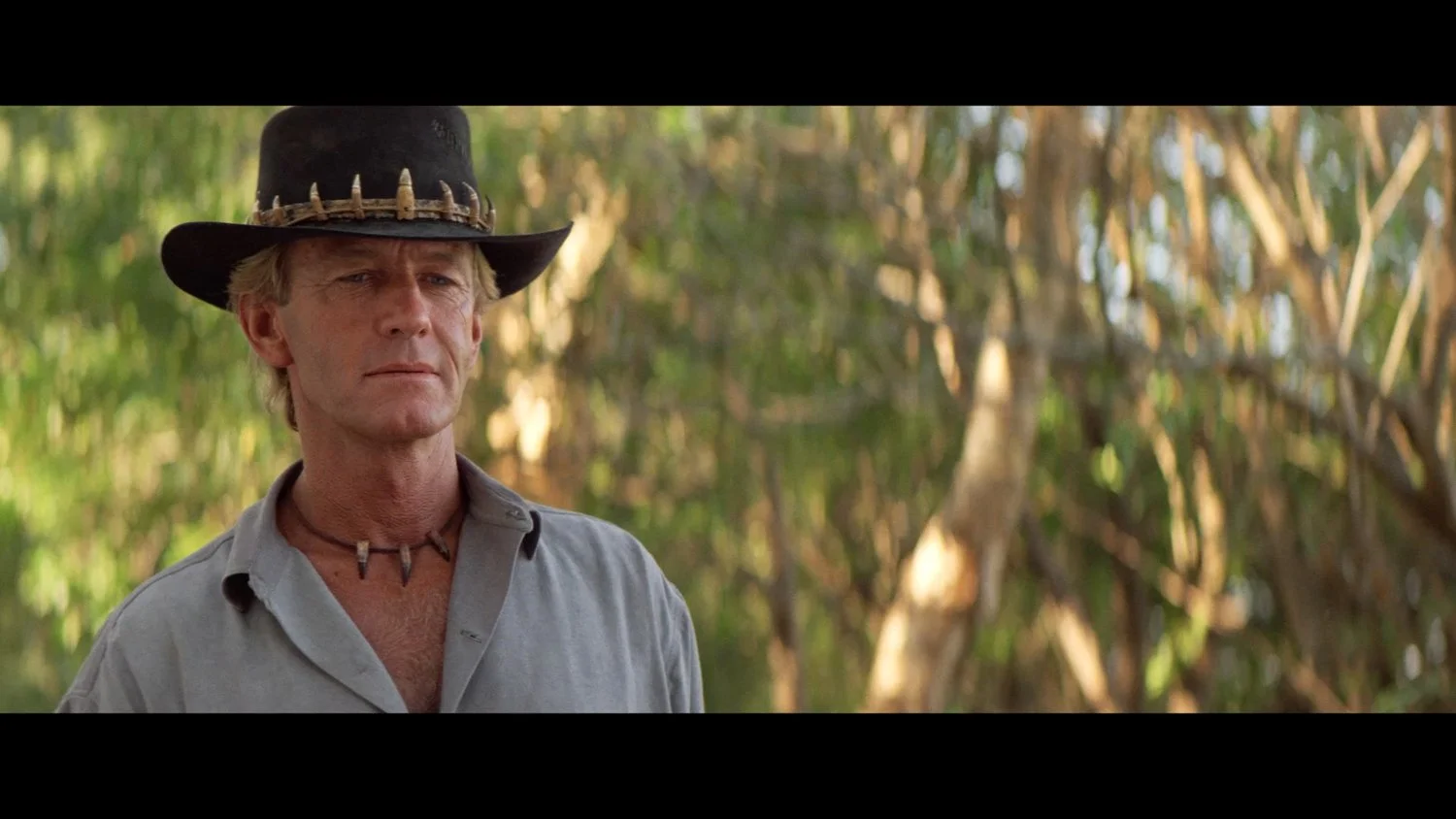 Actor Paul Hogan plays Mick Dundee wearing a khaki shirt and a black hat with teeth decorations, standing outdoors in the Australian outback.