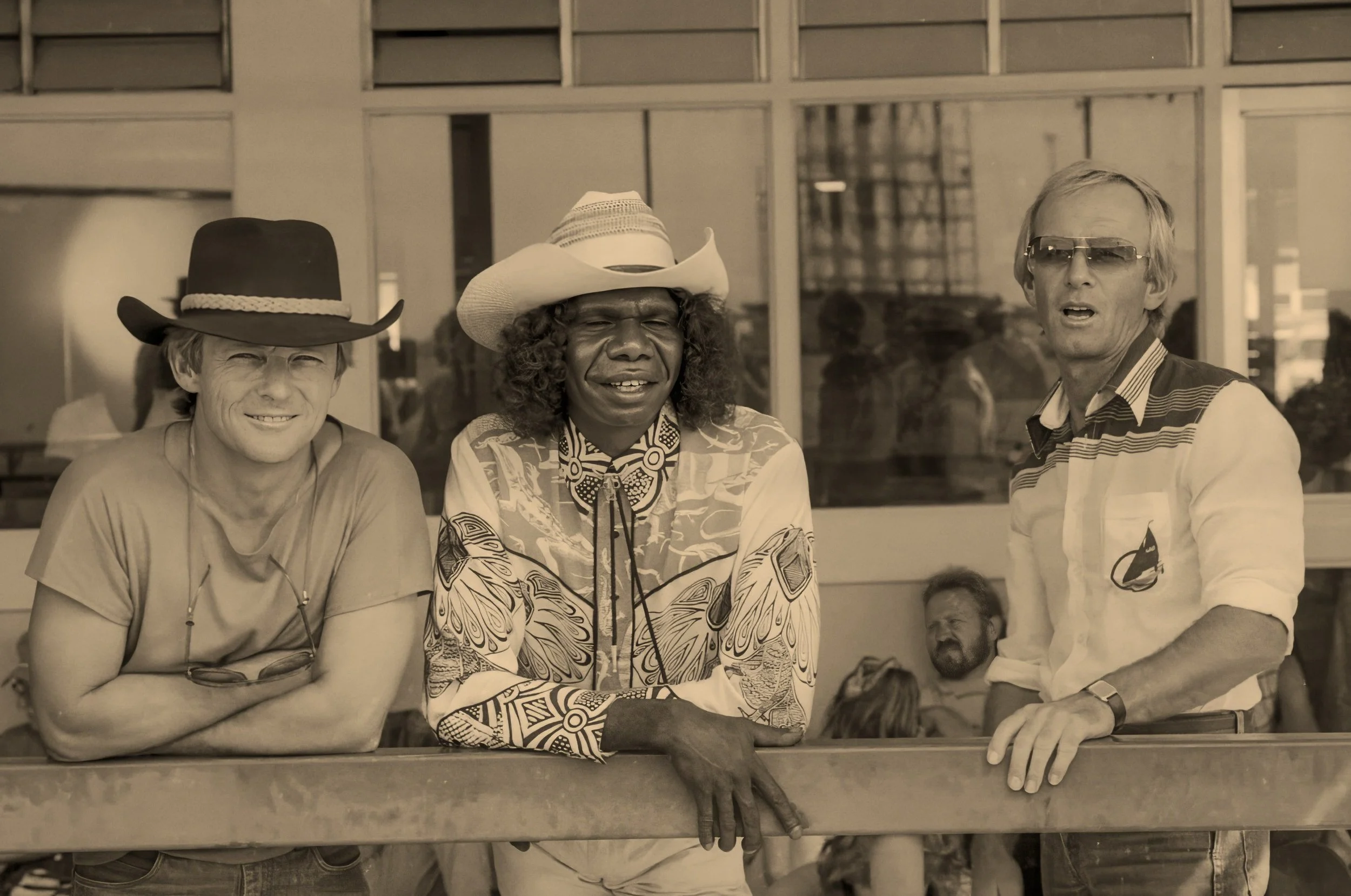 John Cornell, David Gulpilil and Paul Hogan standing together outside a building, smiling and talking, with a reflection of others in the glass behind them.