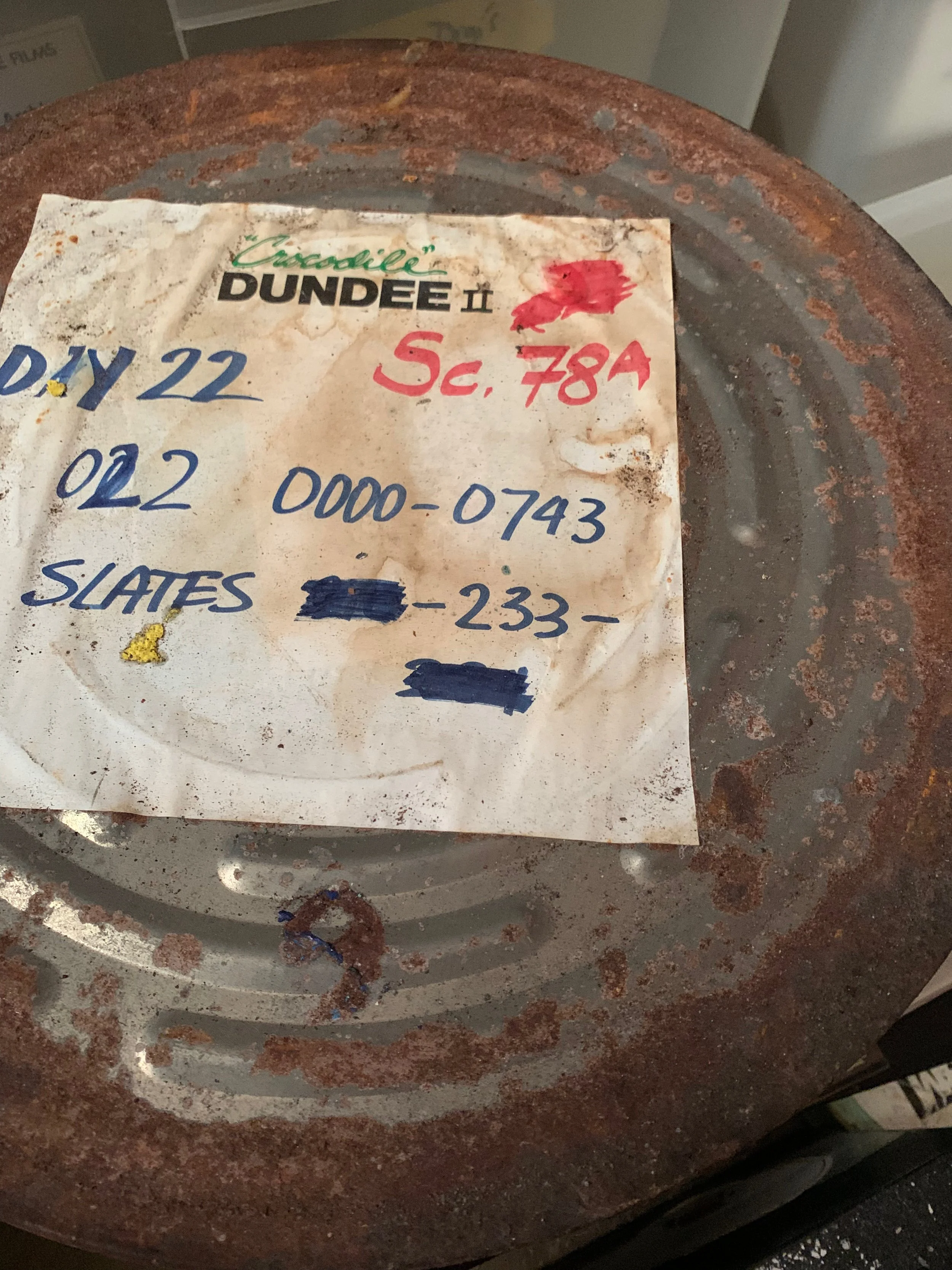 Rusty metal surface with a worn, stained handwritten note on it, listing a date, location, a phone number, and partially obscured text.