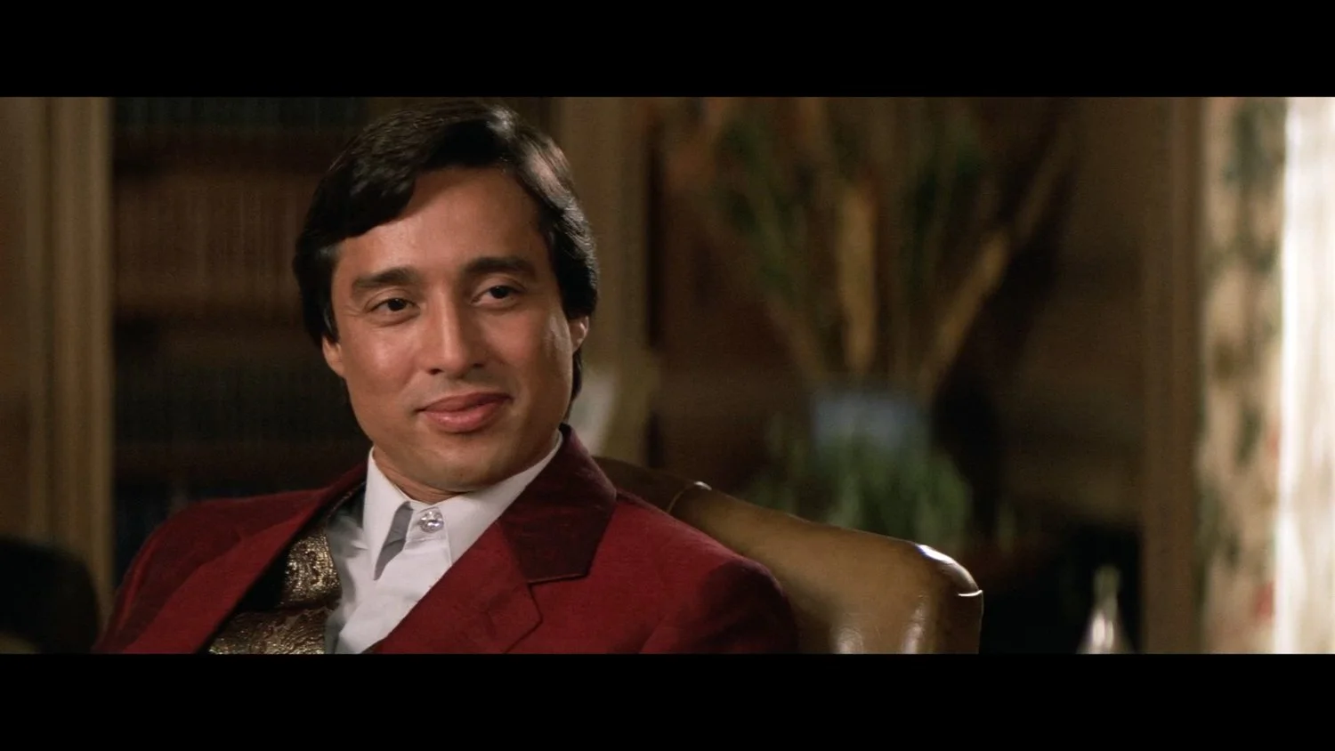 Close-up of actor Hechter Ubarry playing Luis Rico dressed in a red jacket and white shirt, sitting on a brown chair in a room with bookshelves and a potted plant.